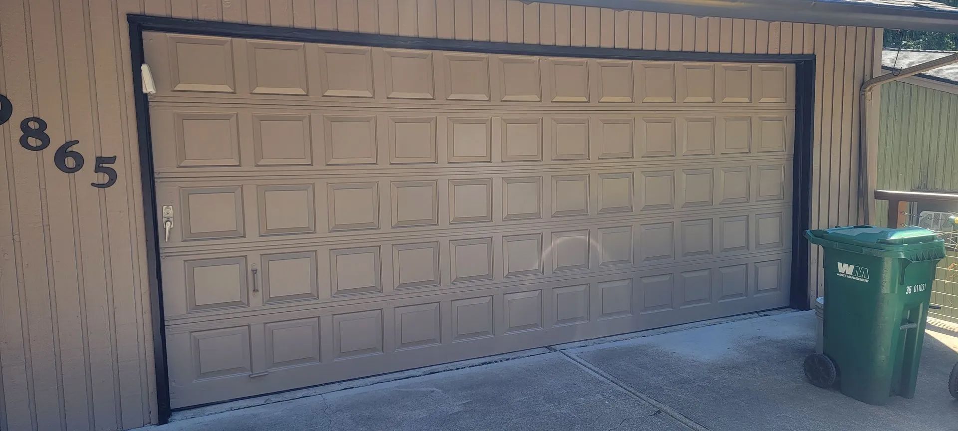 Beige garage door on a house, with a green trash bin beside it and house number 865 on the wall.