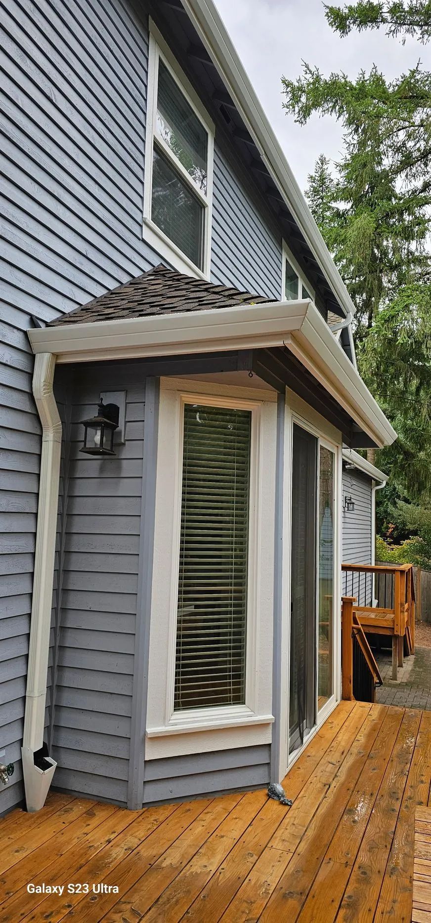 Small gray house extension with bay windows and a shingled roof on a wooden deck, beside blue siding and trees.