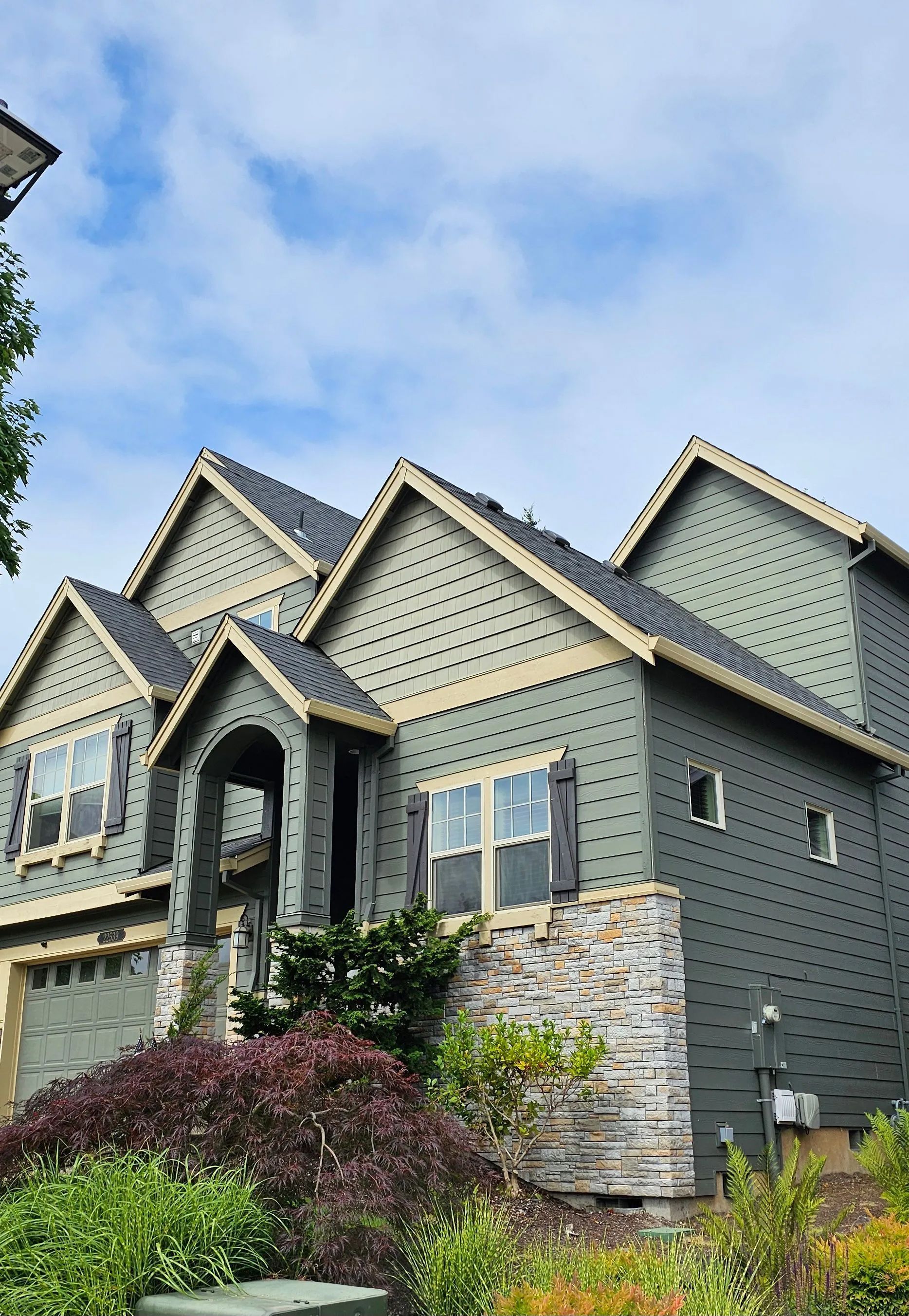 Suburban house with gray siding and stone accents, gabled roofs, and landscaped shrubs under a blue sky