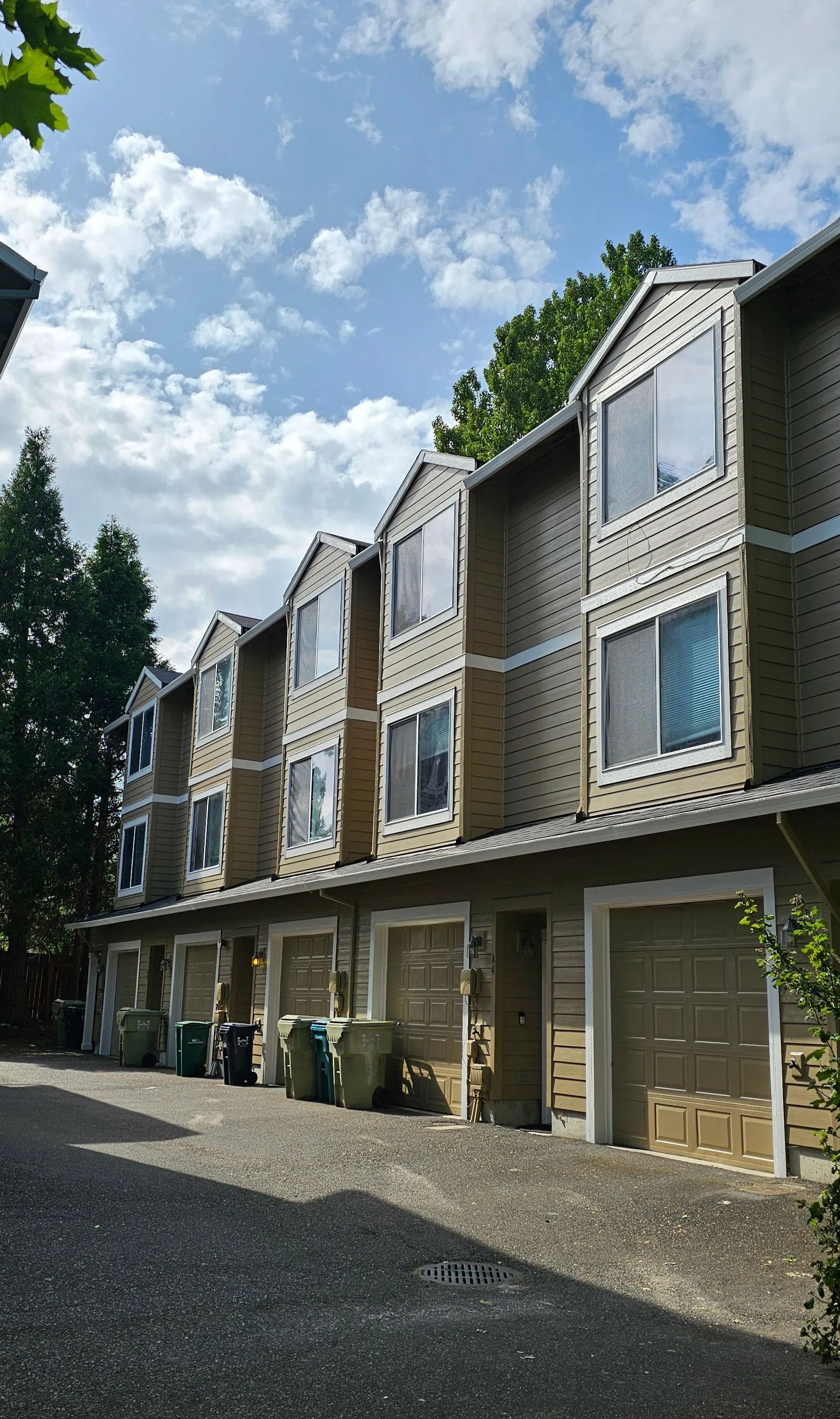 Row of beige townhouses with garages on a sunny street under a partly cloudy sky