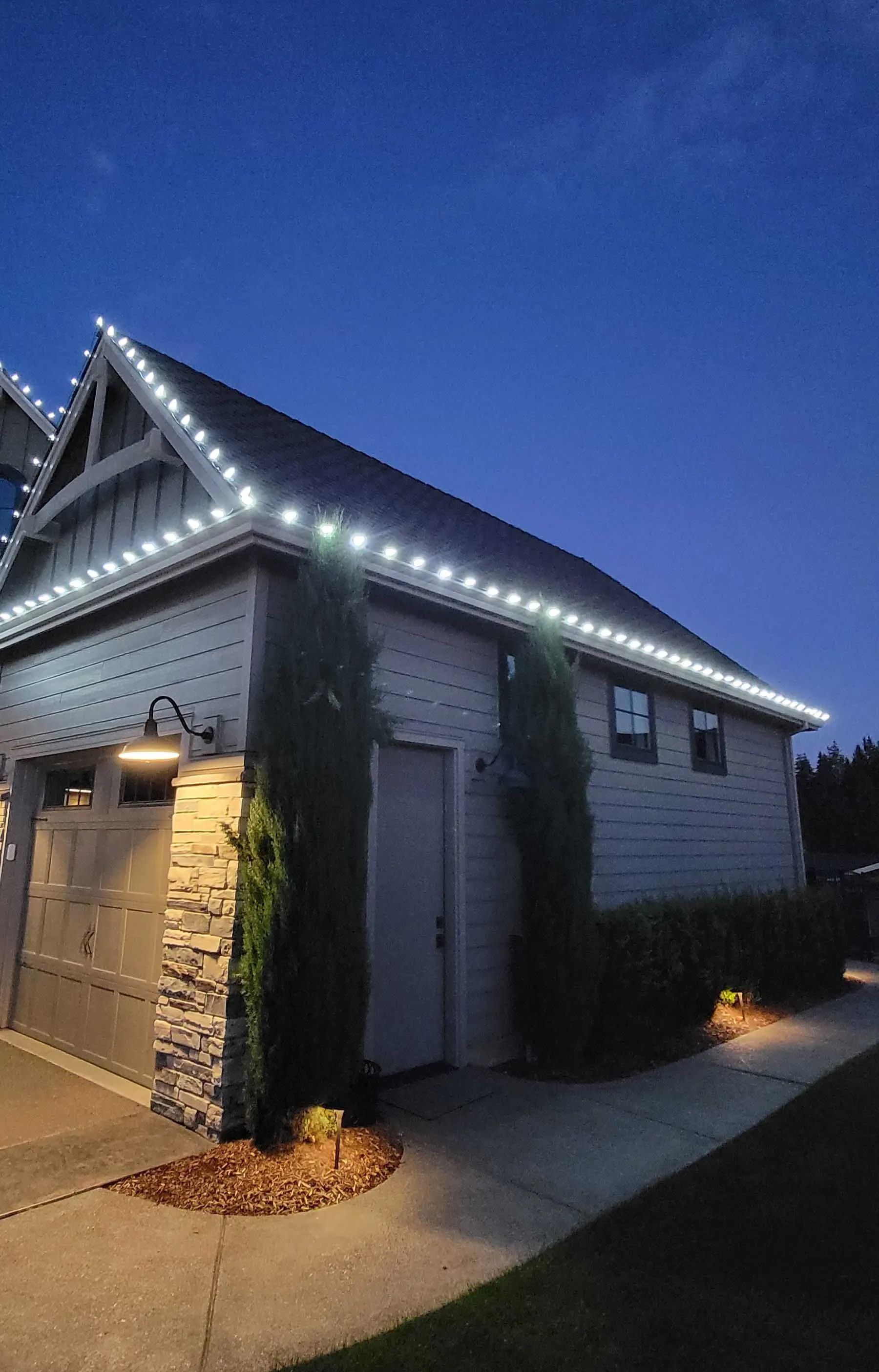 Two-story gray house with roofline lights at dusk, lit garage and walkway below a blue sky