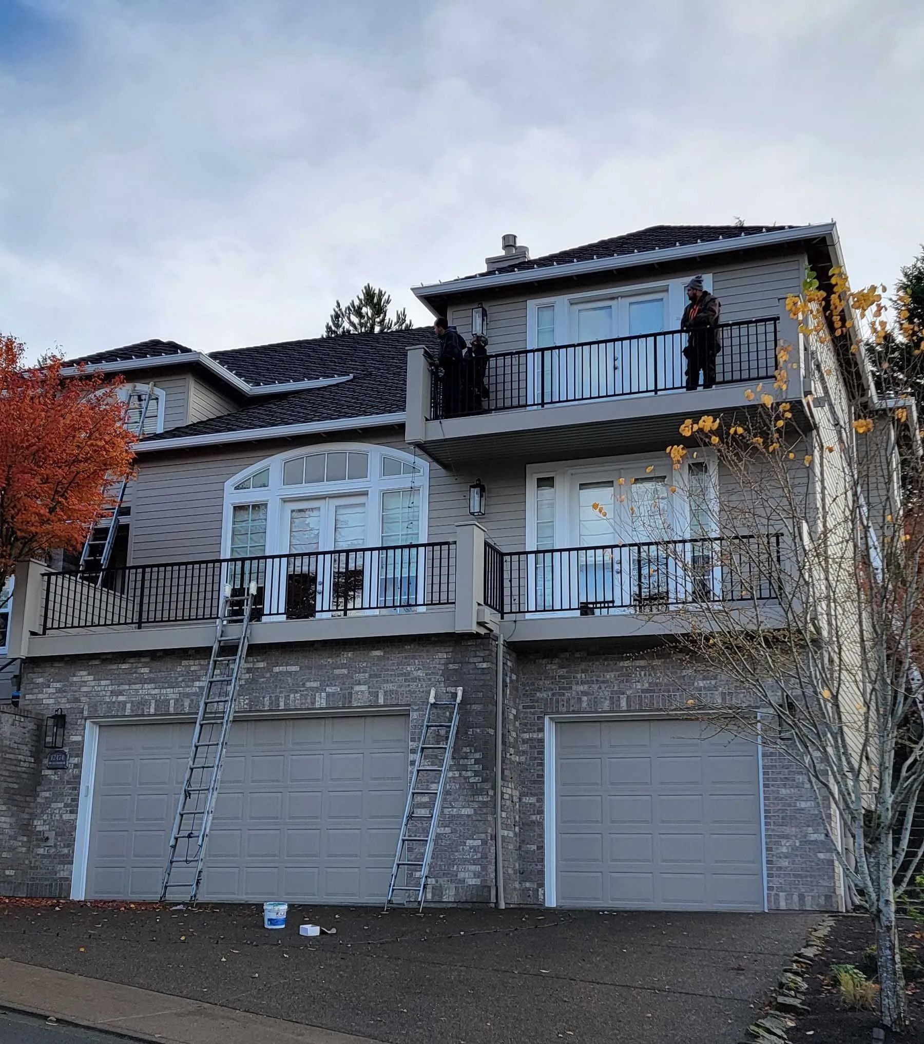 Two-story house with ladders leaning against the garage, balcony, and a person standing on the upper deck.