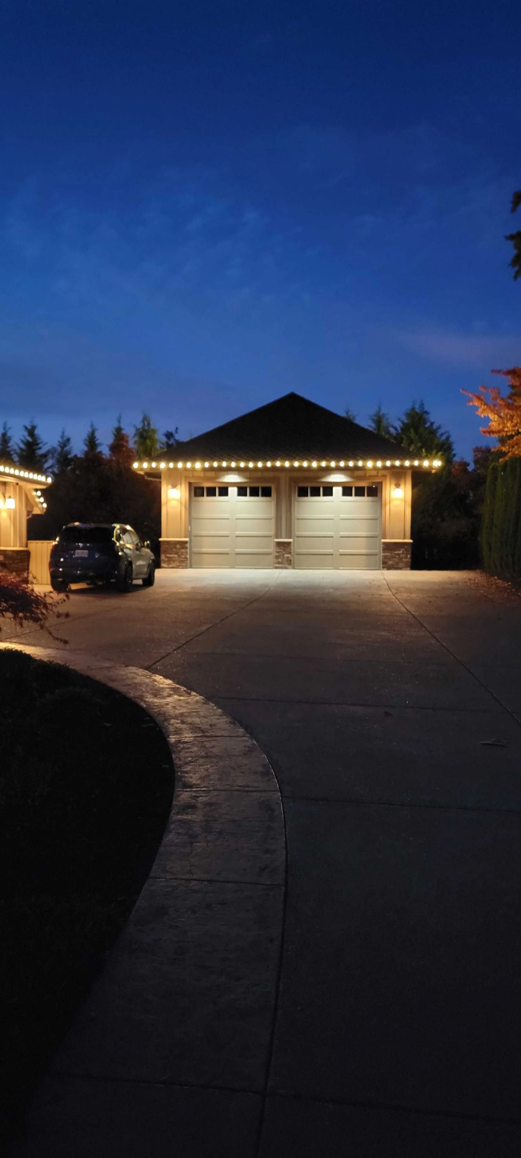 A suburban house at dusk with lit garage lights and a curved driveway under a deep blue sky.