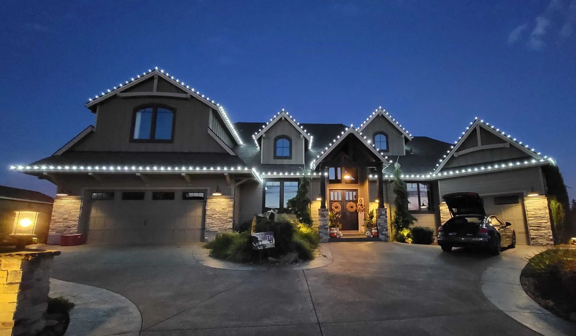 Large illuminated suburban house at dusk with a three-car garage and driveway, lights glowing warmly inside