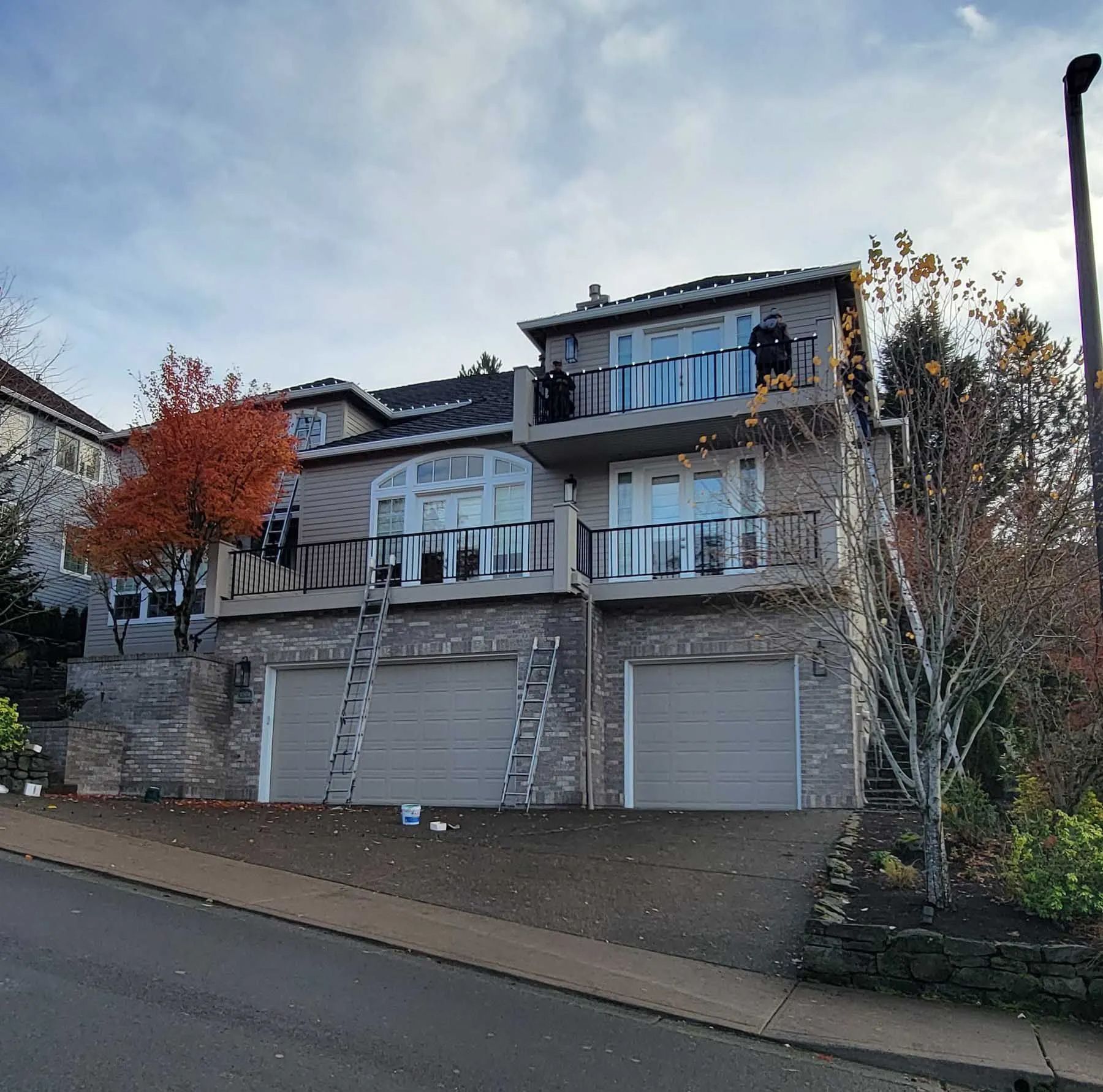 Large beige two-story house with balconies, stone retaining wall, garage, and autumn trees by the street