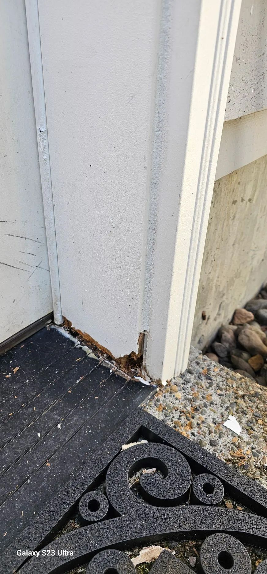 Close-up of a white door frame with peeling paint and rusted base beside a black doormat and gravel edge