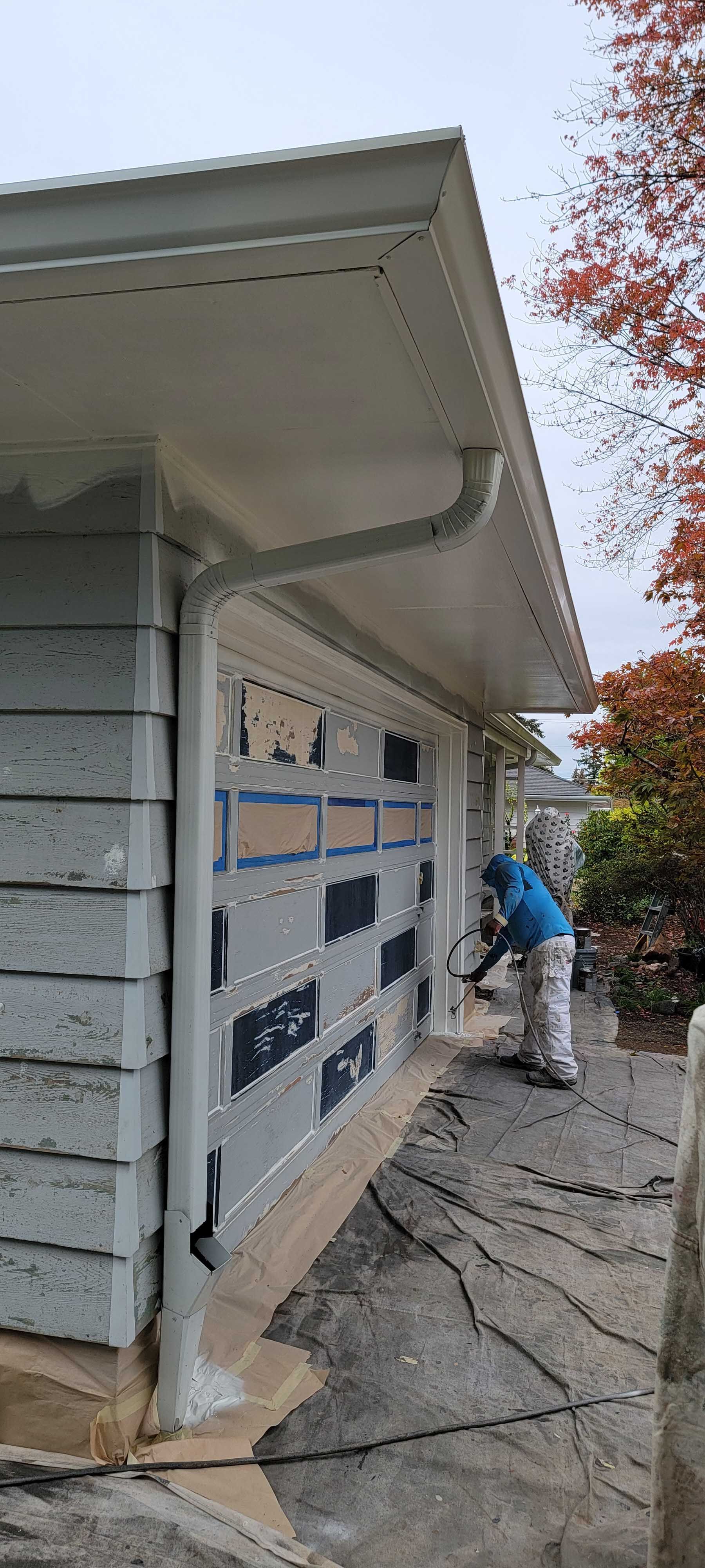 Person painting a gray garage door, white trim, with a gray house and roof. Overcast day.