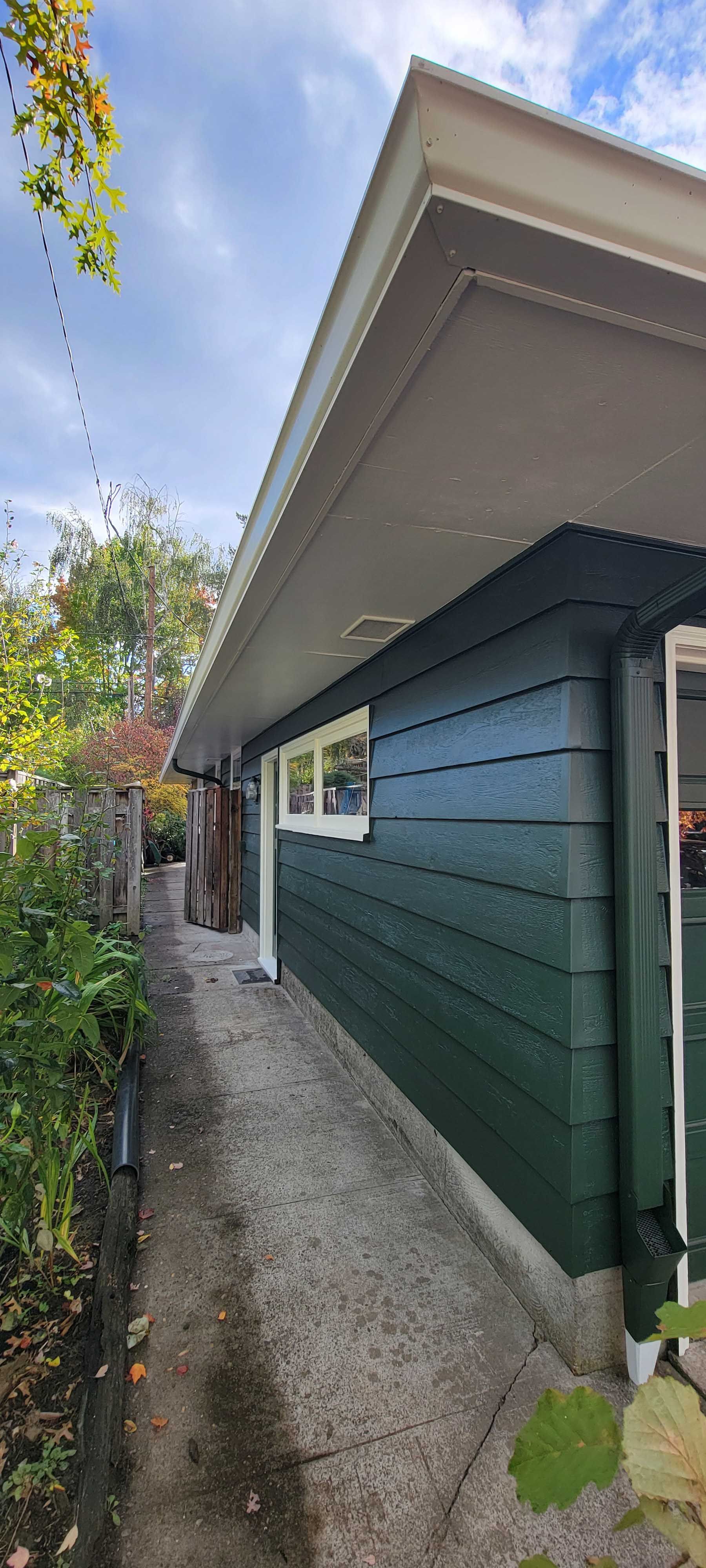 Side view of a green building with white trim, a concrete path, and foliage under a cloudy sky.