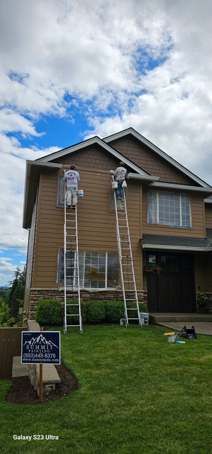 Two workers painting a two-story brown house from ladders. The sky is partly cloudy and the grass is green.