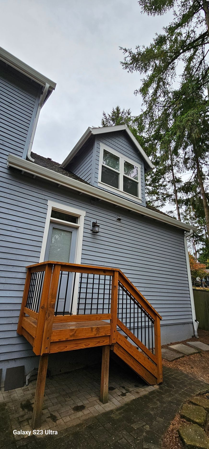 Back exterior of a blue house with a wooden deck and dormer windows, under a cloudy sky.