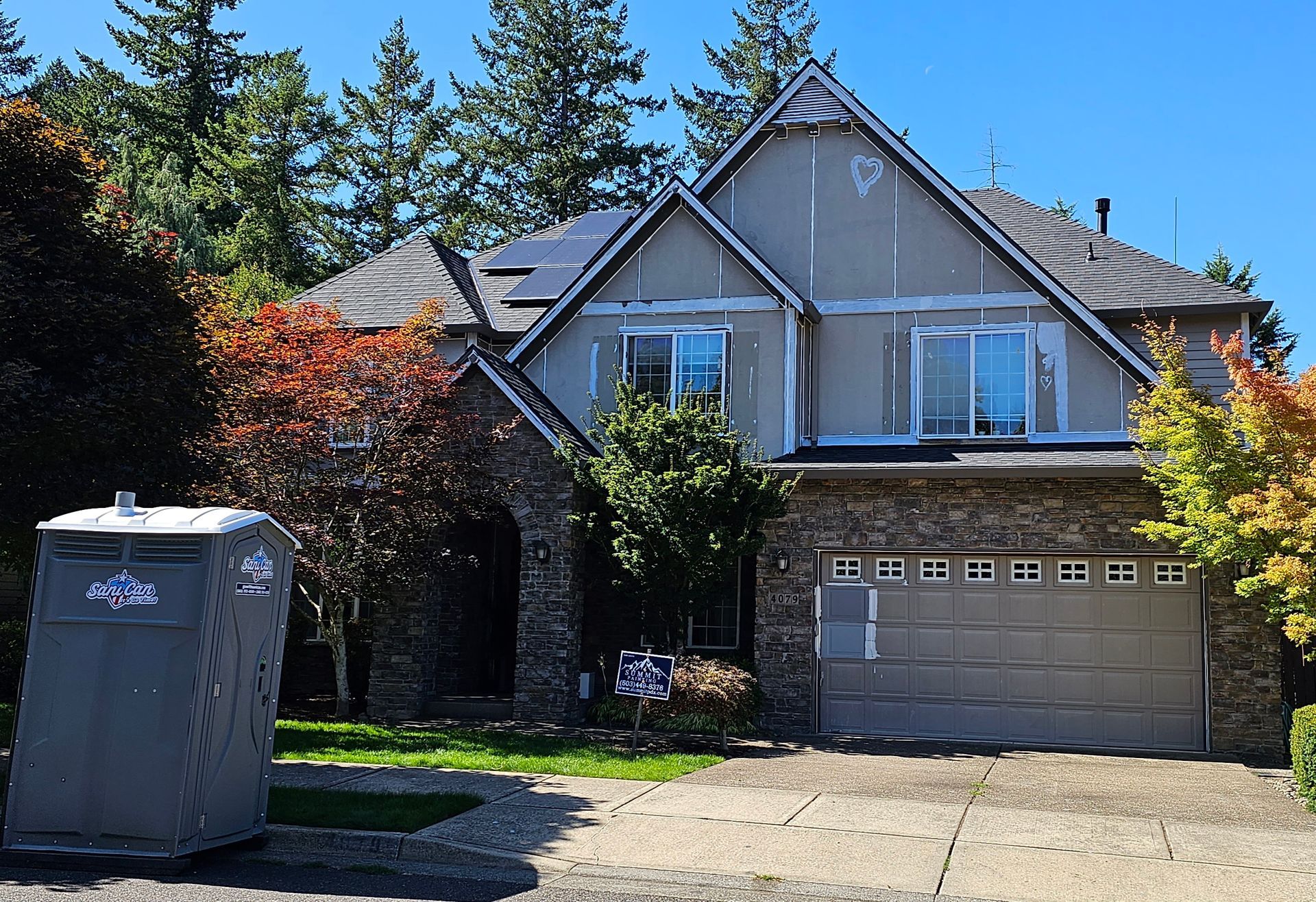 A large house with a gray garage door, stone accents, and a porta-potty in the yard.