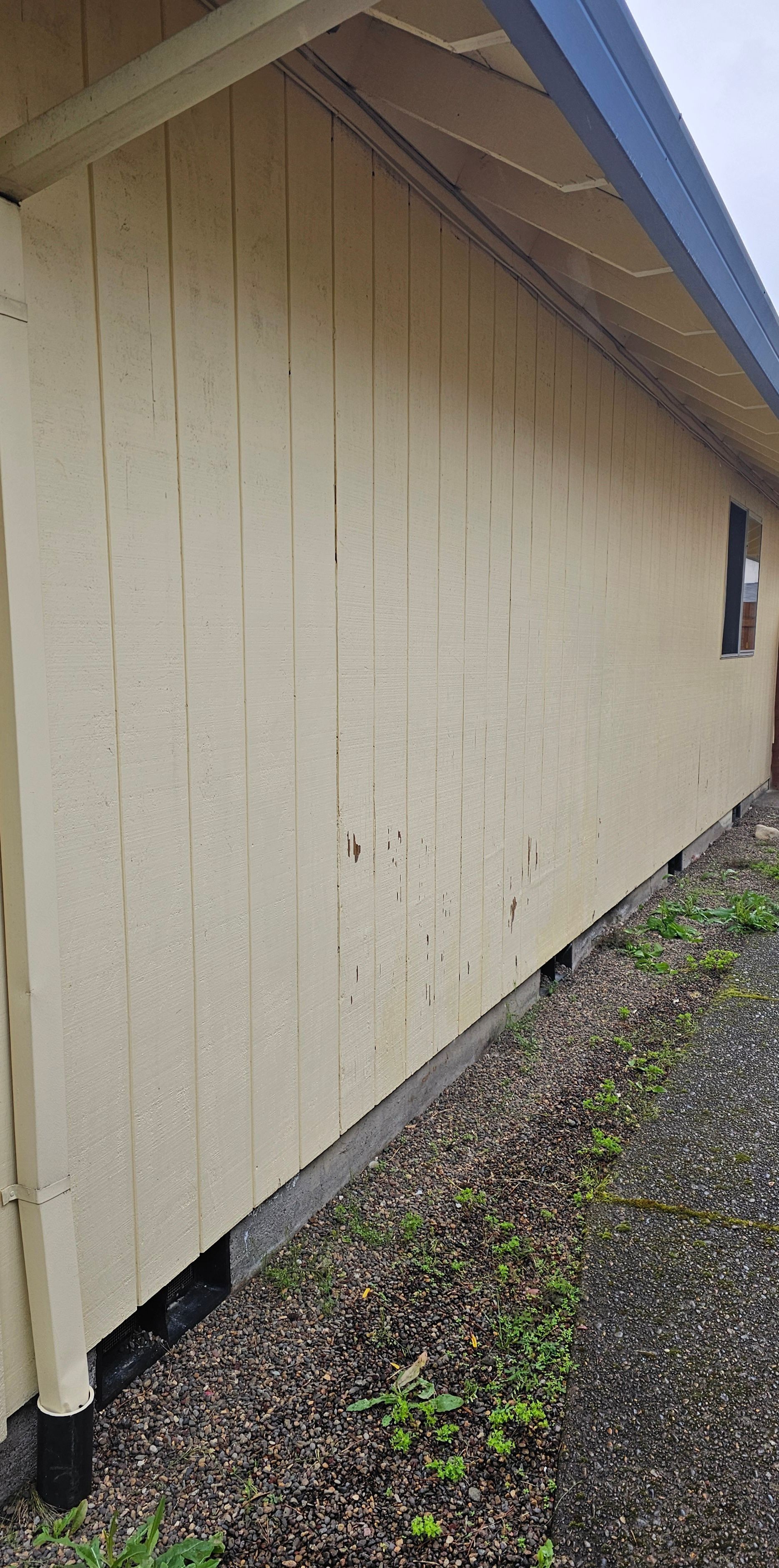 Light yellow wooden exterior of a building with vertical planks. Black roofline and gravel ground.