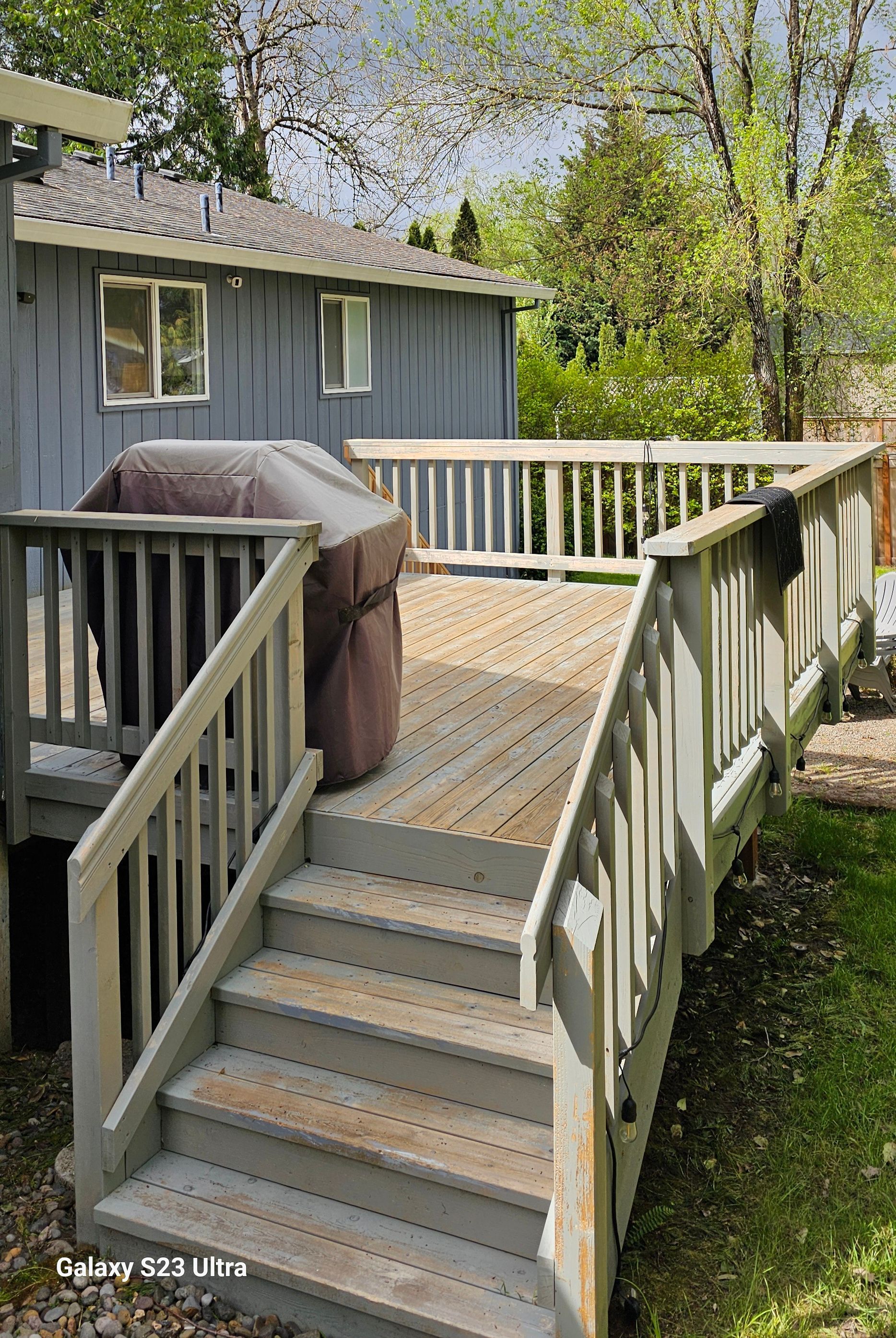 Gray wooden deck with stairs and grill, next to a gray building and trees.