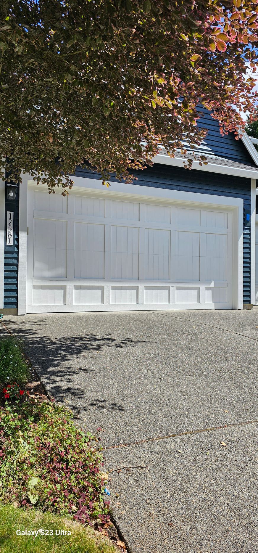 White garage door on a house with a gravel driveway and tree, under a blue sky.