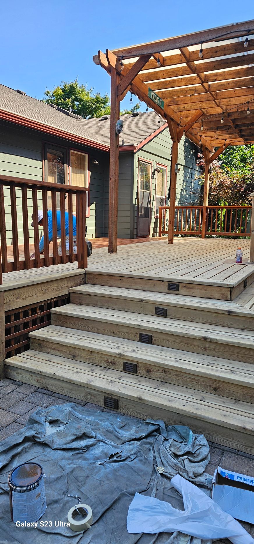 Wooden deck with steps, a pergola, and work items on a tarp; exterior view.