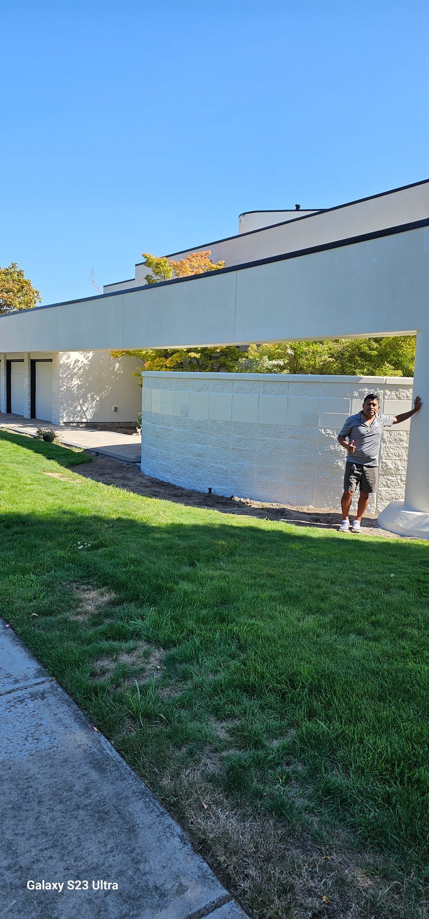 A person stands near a white building with a curved wall. Green grass and blue sky.