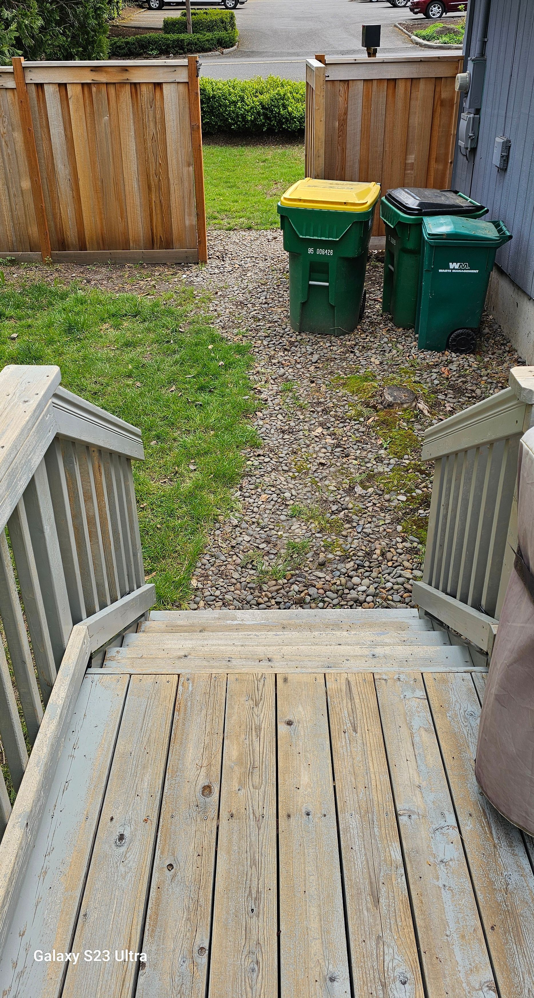 Wooden stairs lead to a small gravel area with green trash cans and a wooden fence.
