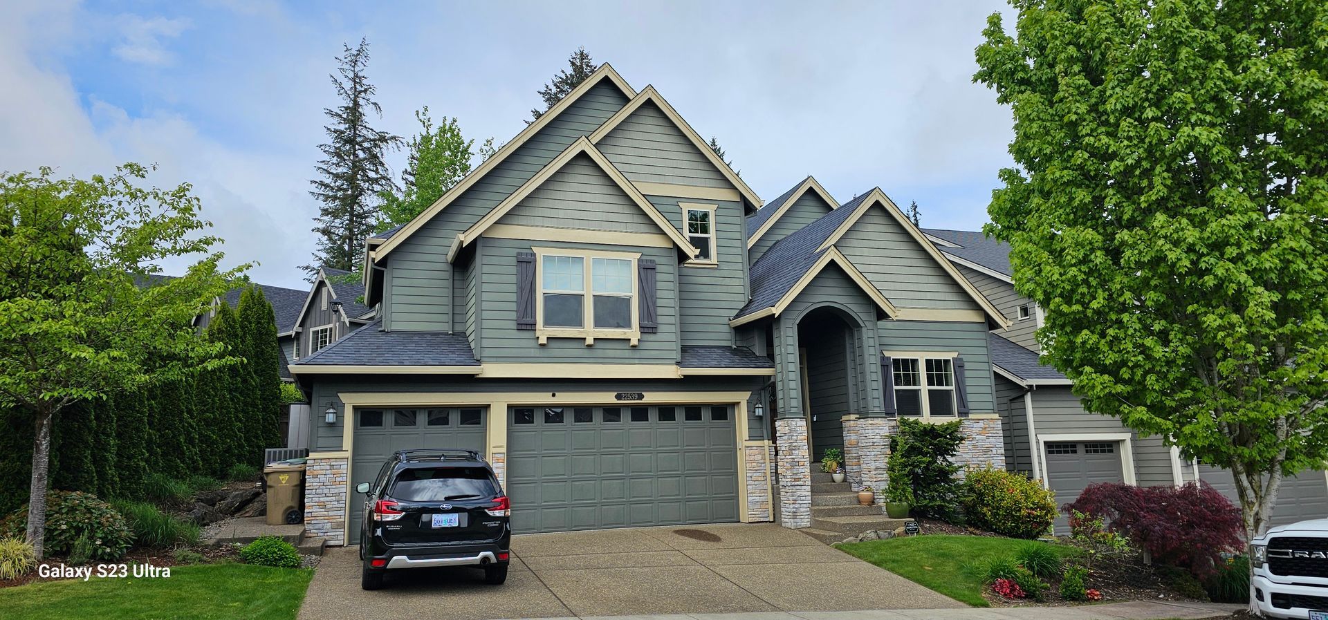 Green house with three-car garage. A black car is parked in the driveway. Trees and green bushes surround the house.