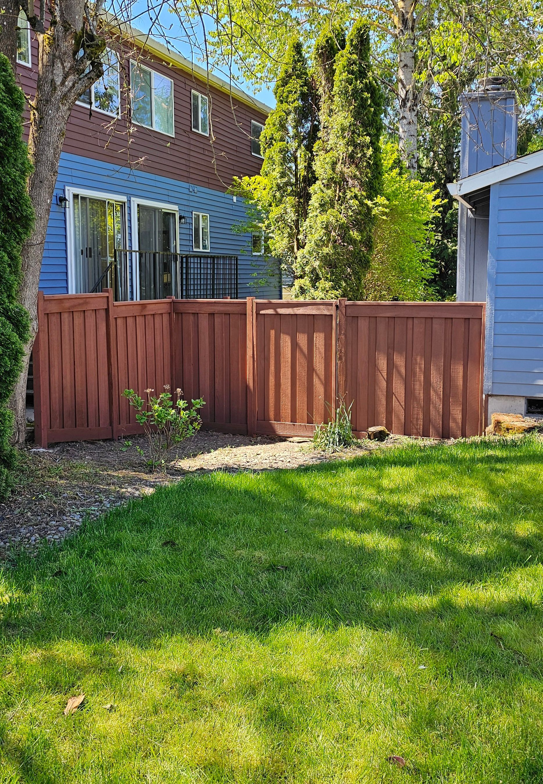 Red fenced backyard with grass, next to a blue house and trees.