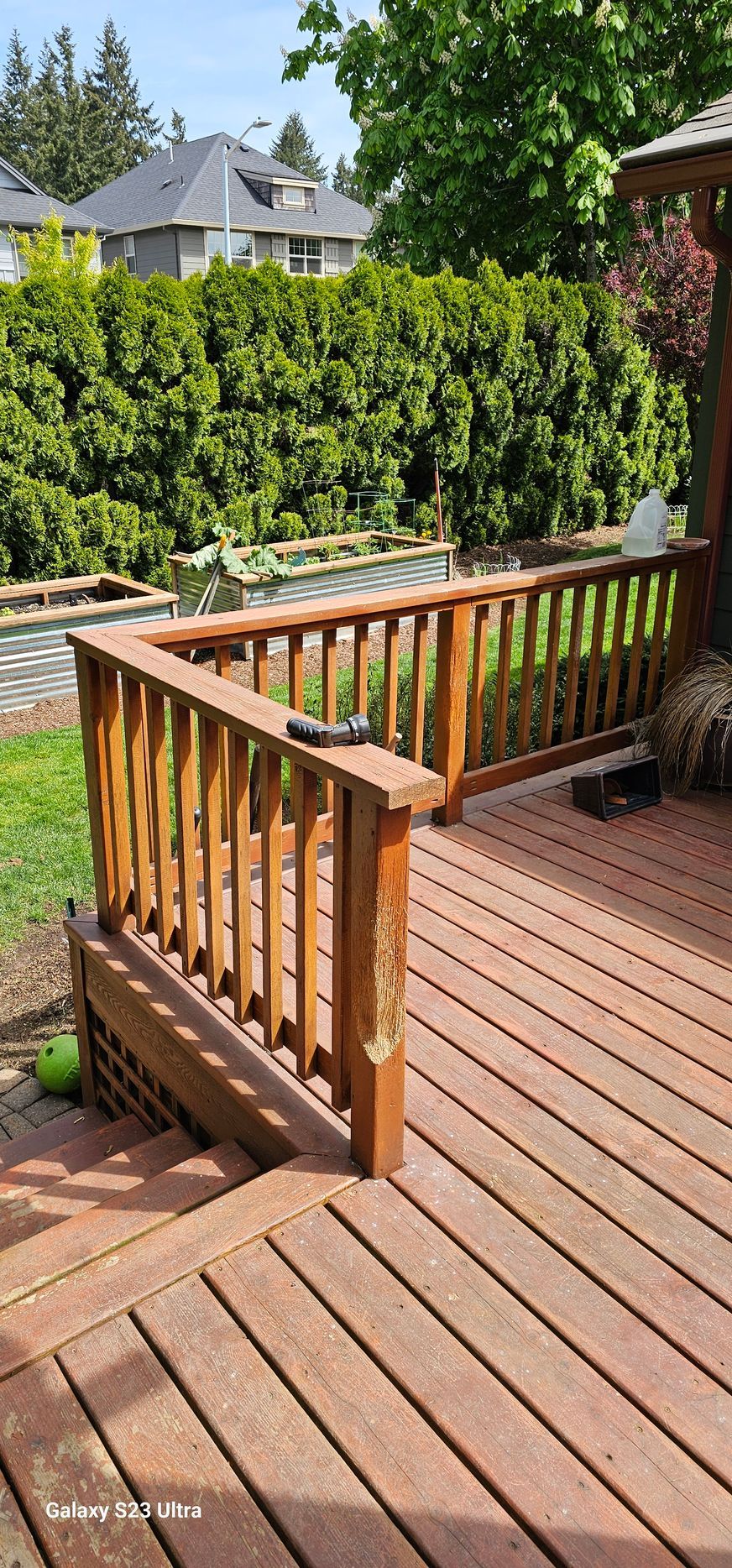 Wooden deck with railing, a green lawn and hedge, and houses in the background.