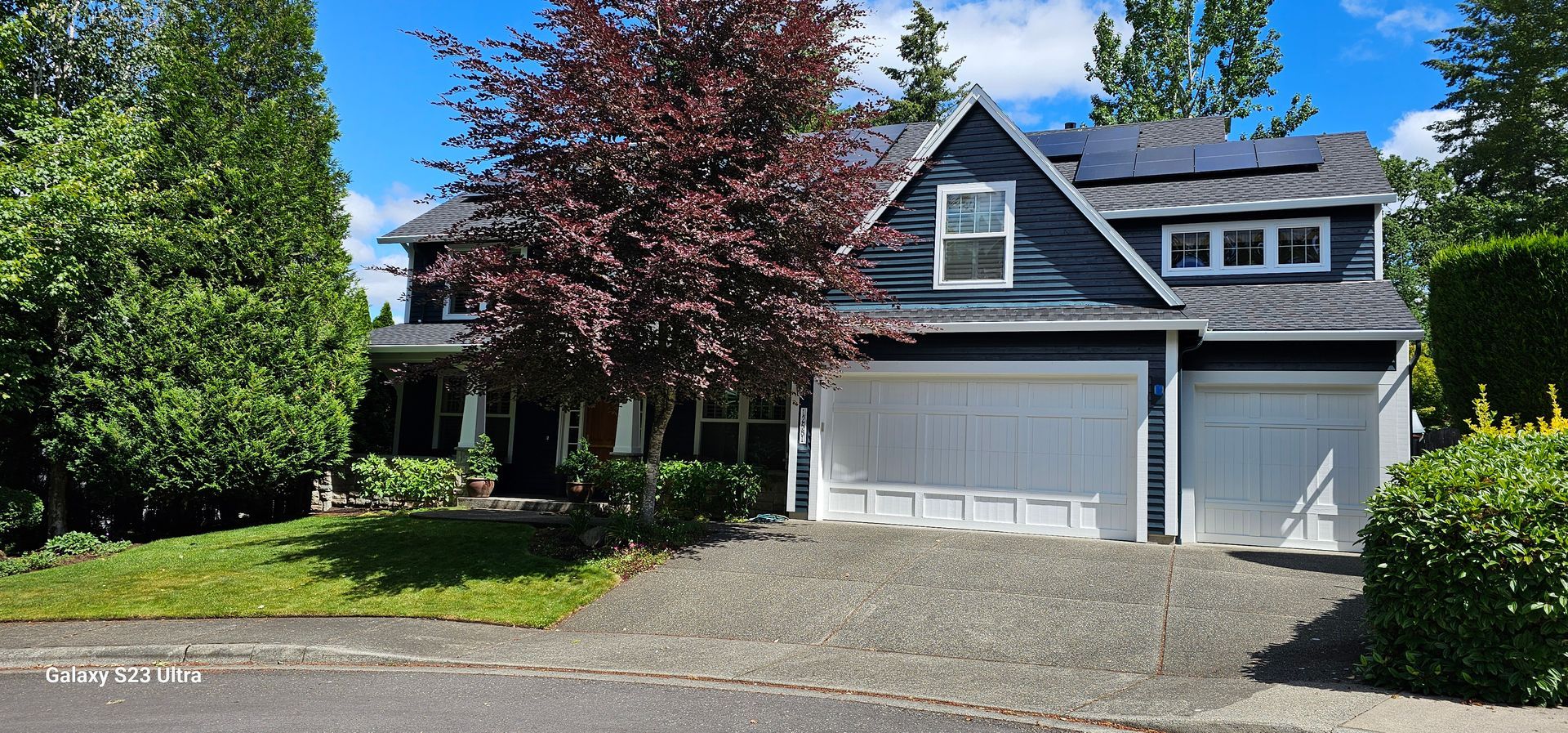 Blue house with white garage doors, red tree, and green lawn on a sunny day.
