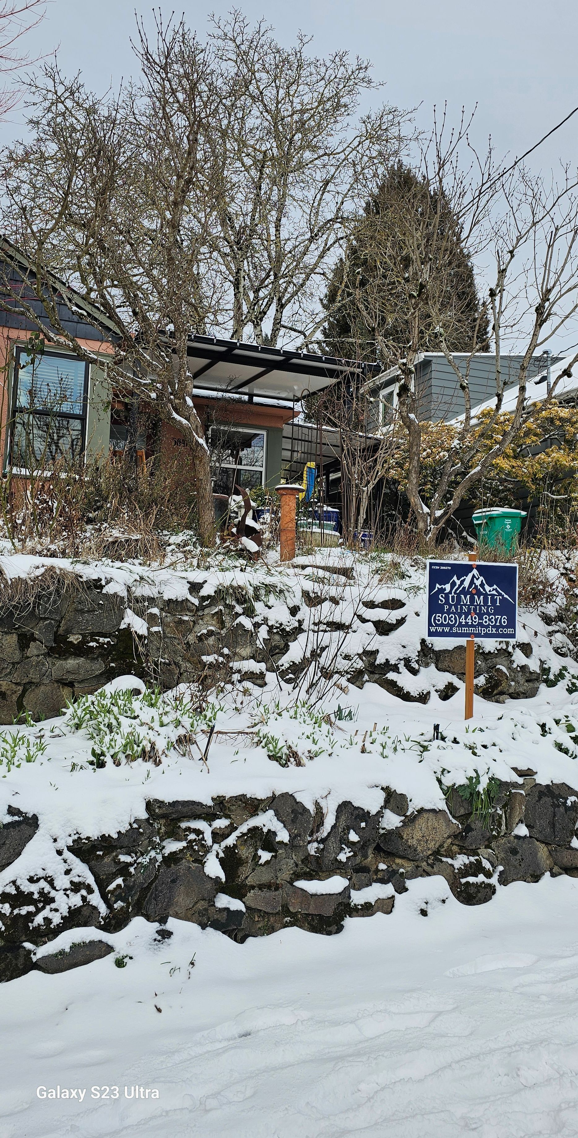 Snow-covered stone wall with a blue sign. A house and trees are visible in the background.