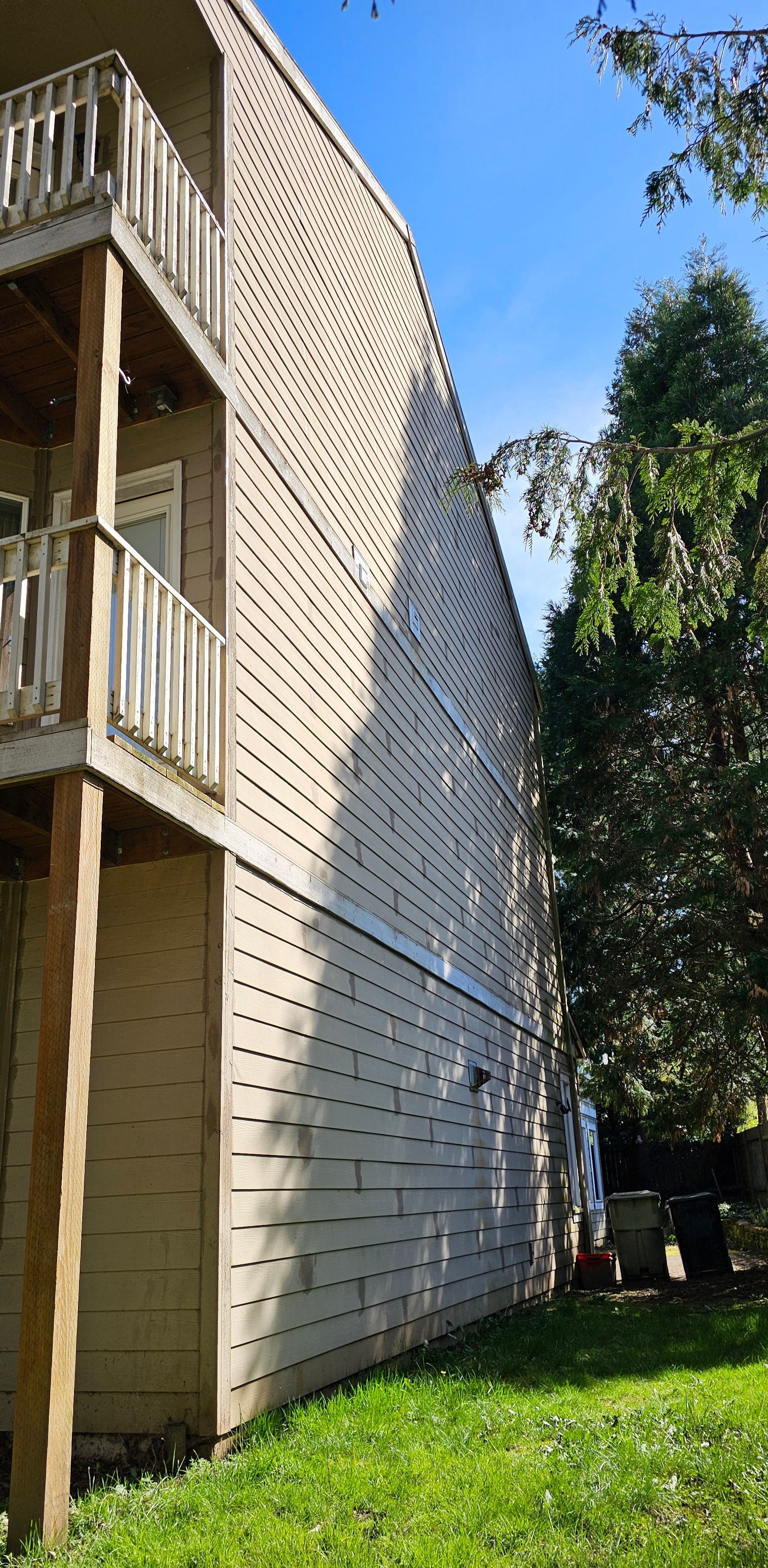 Tan apartment building with balconies, next to a green lawn and trees, against a blue sky.