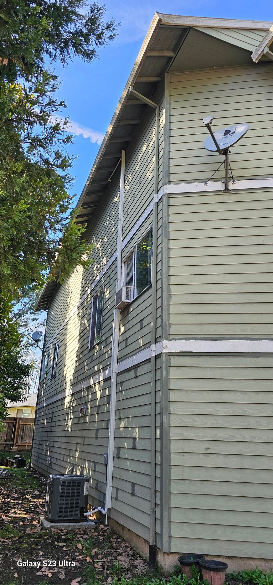 Side of a two-story light green building with satellite dish, windows, and white trim. Blue sky and tree visible.