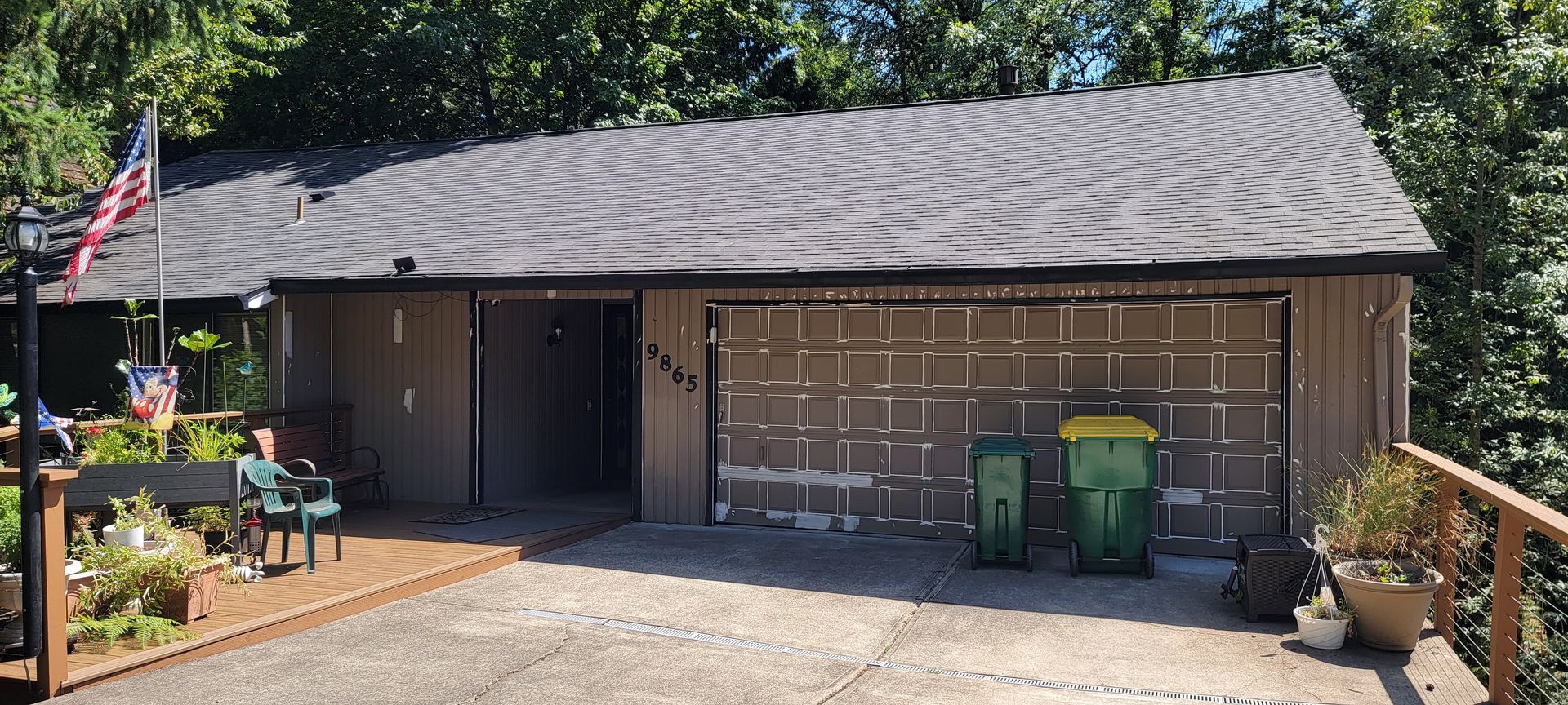 Brown house with garage, American flag, and trash cans. Wooden deck and lush trees surround the structure.