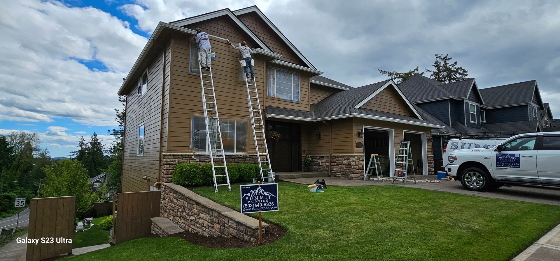 Two workers on ladders painting a two-story house with green grass and a white truck parked in front.