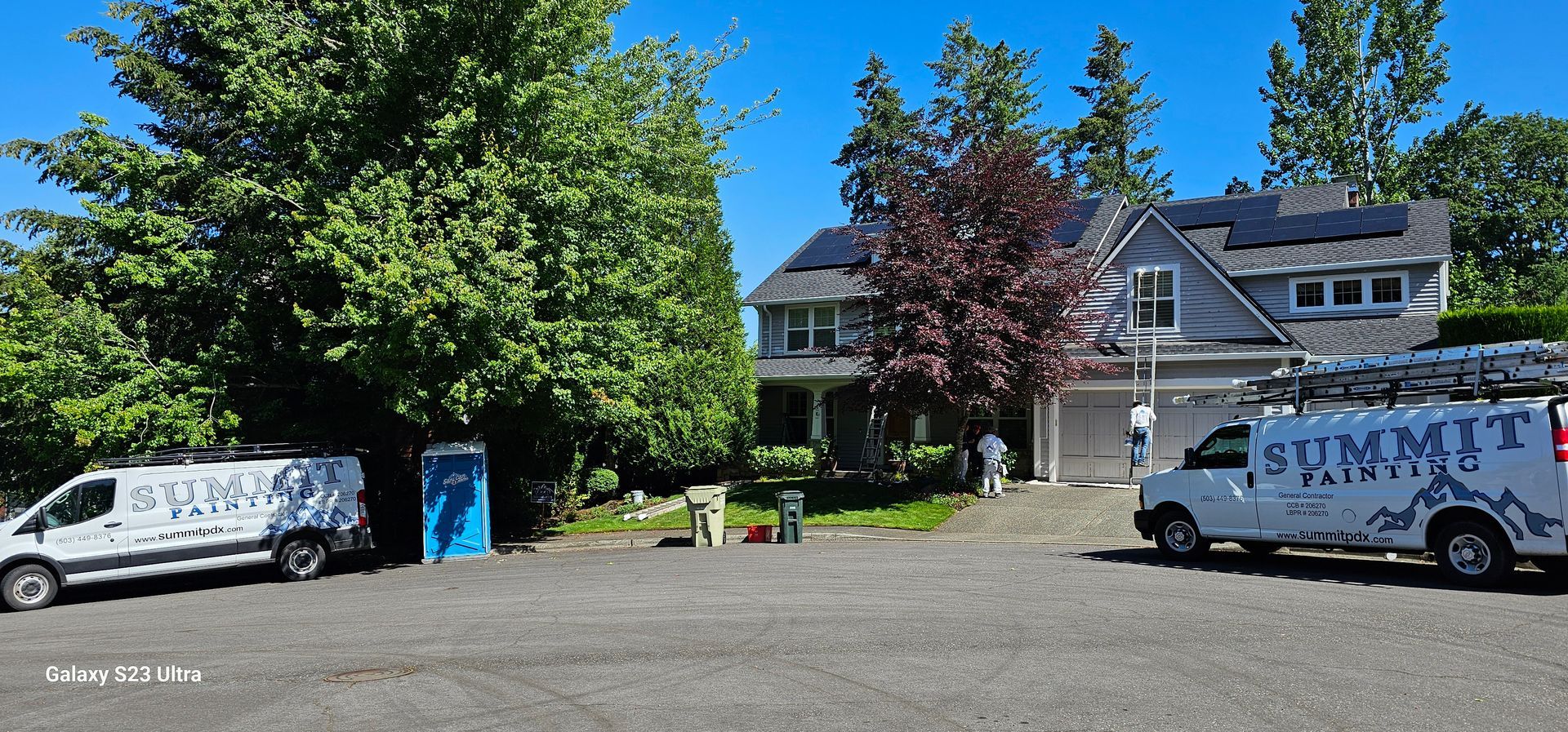 Two white vans with logos parked in front of a gray house with solar panels. A blue porta potty is nearby.
