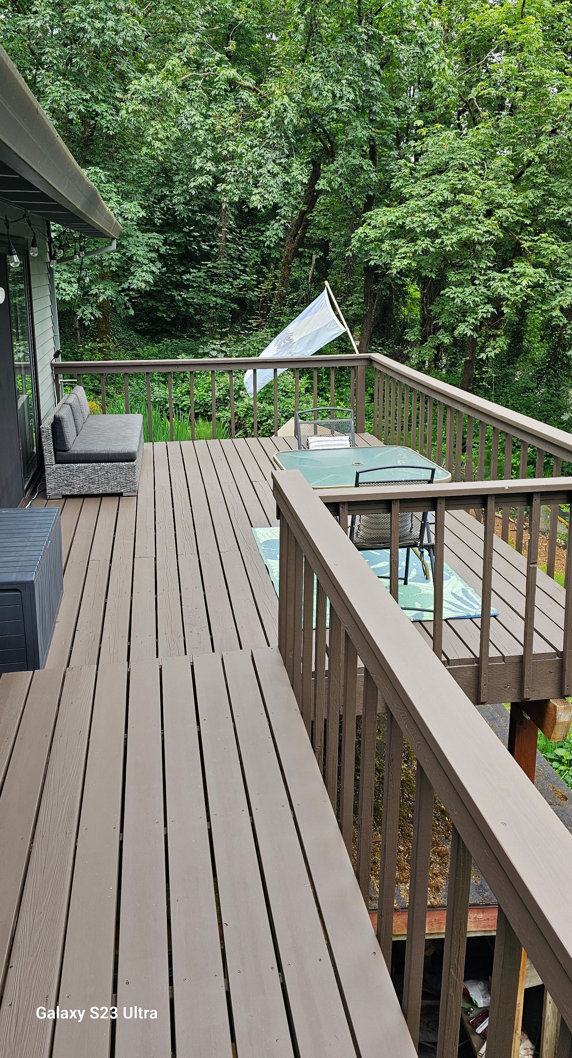 Wooden deck with brown railings, overlooking lush green foliage. Outdoor seating visible.