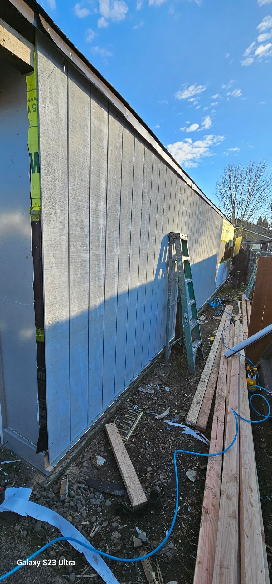 Construction site with gray siding, ladder, lumber, and blue sky.
