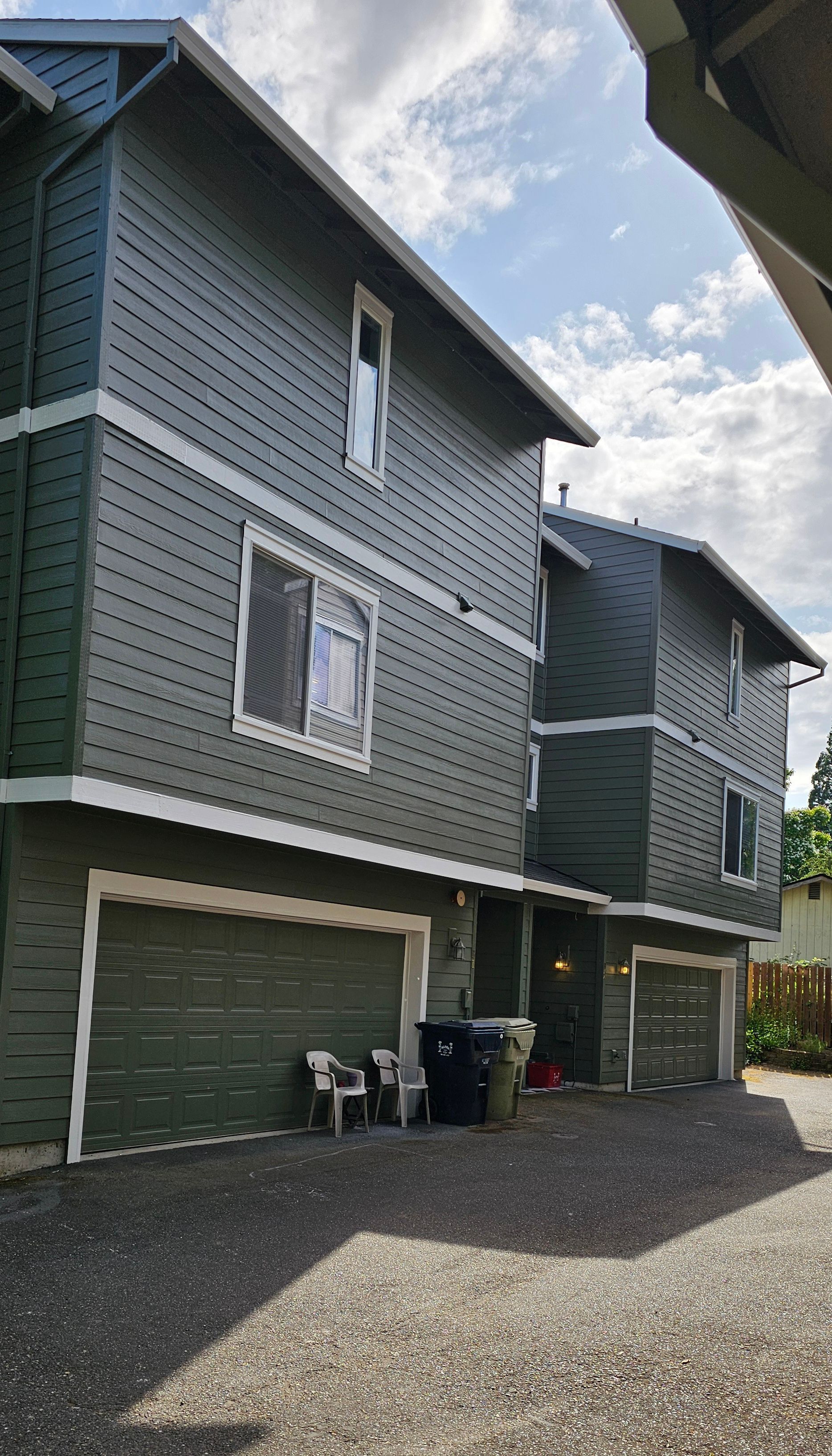 Three-story townhouse exterior with green siding and garage doors. Gravel driveway, cloudy sky.