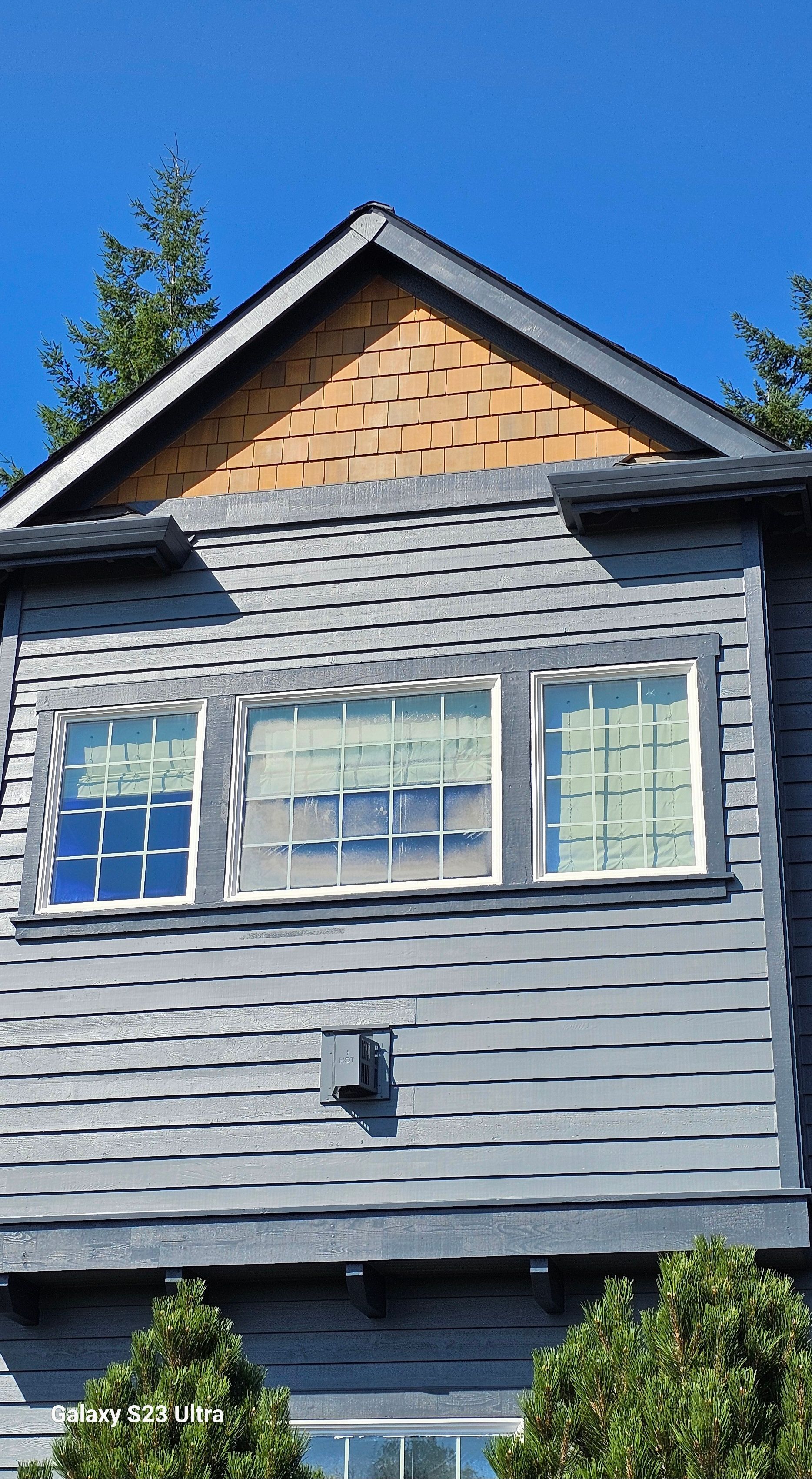 Gray house with three windows and brown roof shingles, under a blue sky.