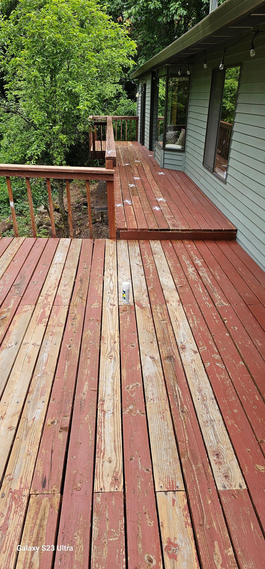 A weathered wooden deck with peeling red paint, adjacent to a house, leads to a lush green treeline.