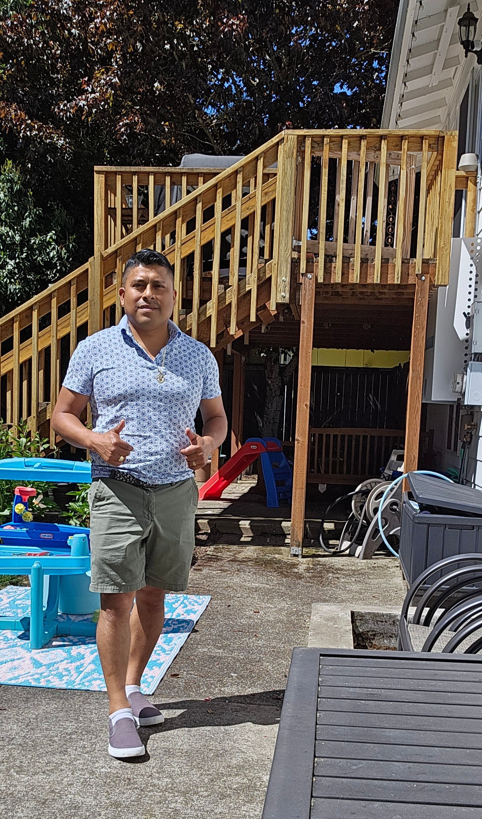 Man in floral shirt and shorts gesturing outdoors near a deck.
