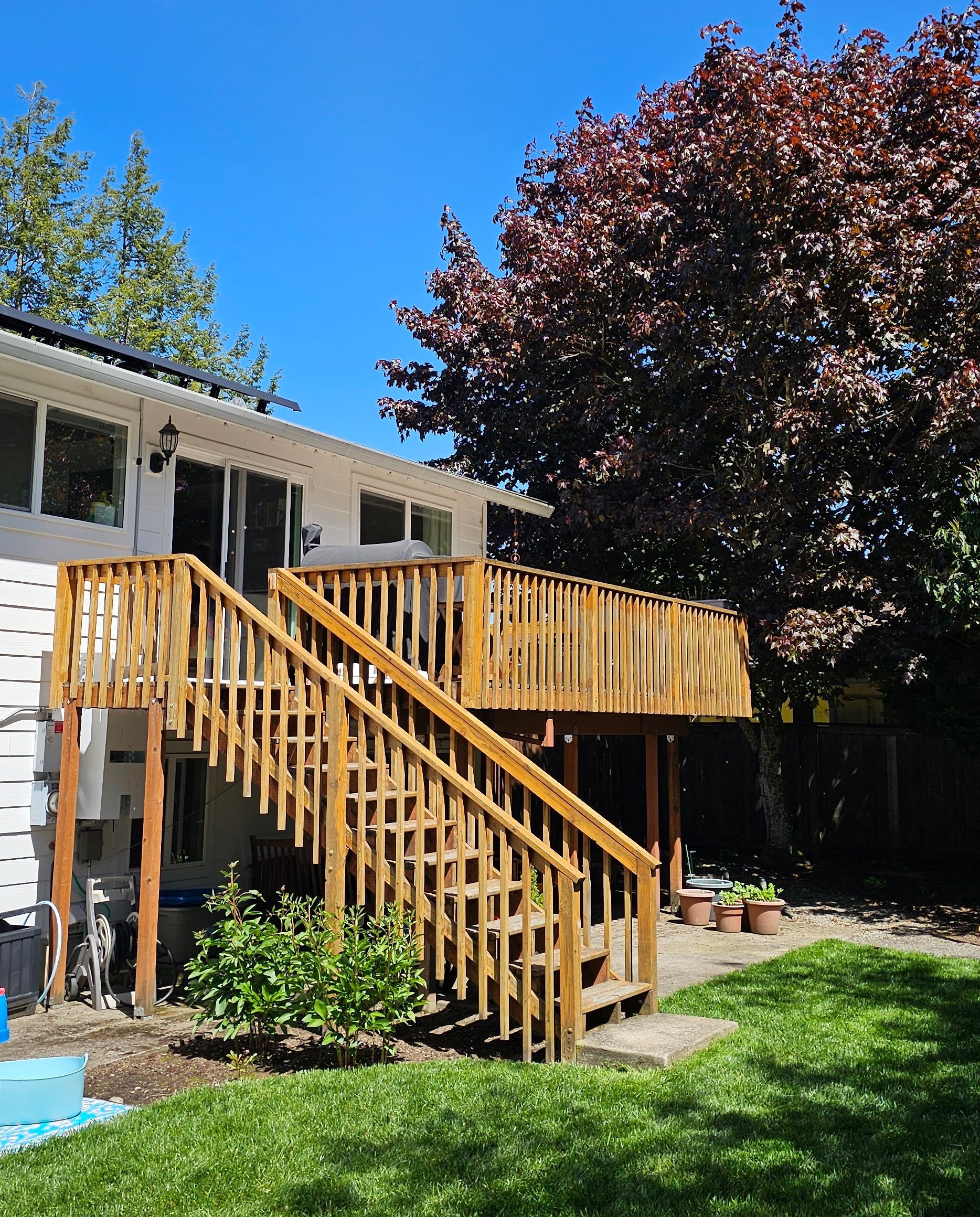 Wooden deck with stairs attached to a two-story white house. Lush green grass and a large tree in the sunny backyard.