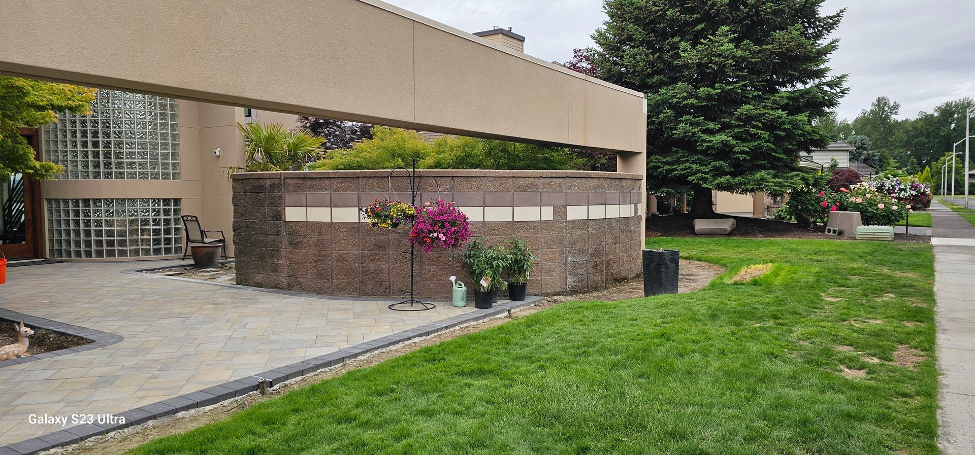 Exterior of a building with a stone wall and lush greenery, a paved area, and a gray sky.