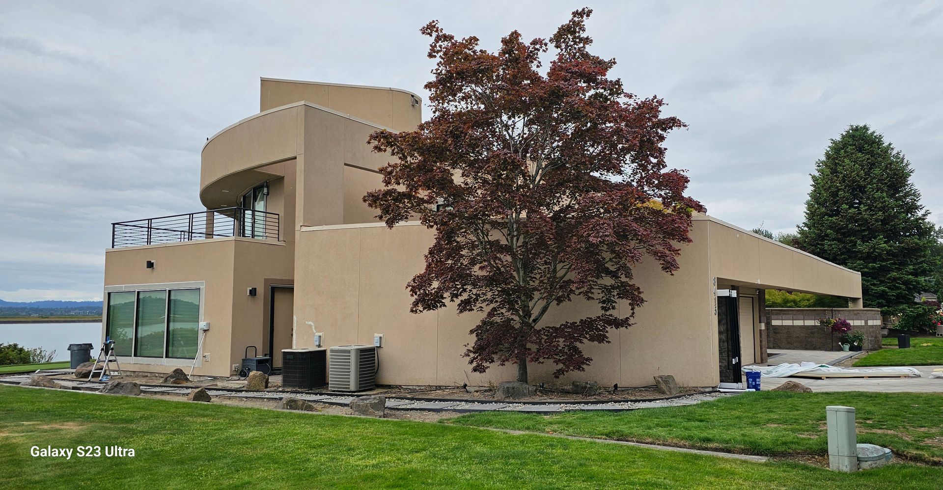 Beige modern home with curved sections, large windows, and a tree with red leaves on a grassy lawn near water.