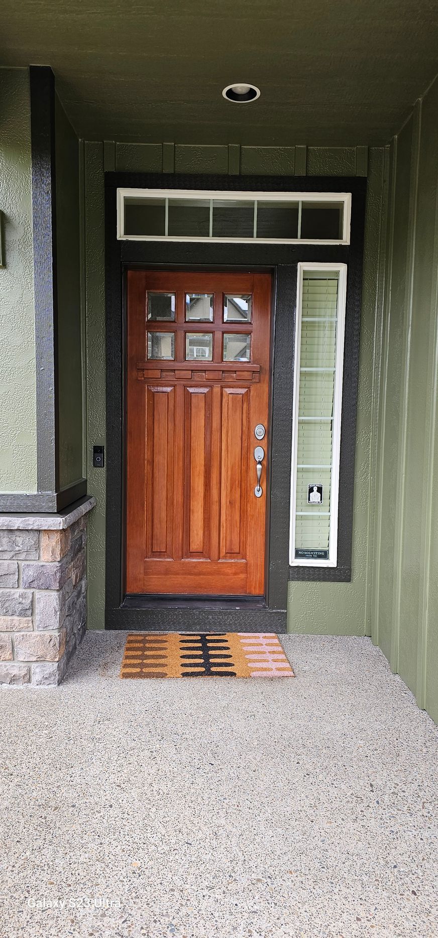 A wooden front door with sidelight and transom, green exterior, and textured concrete walkway.