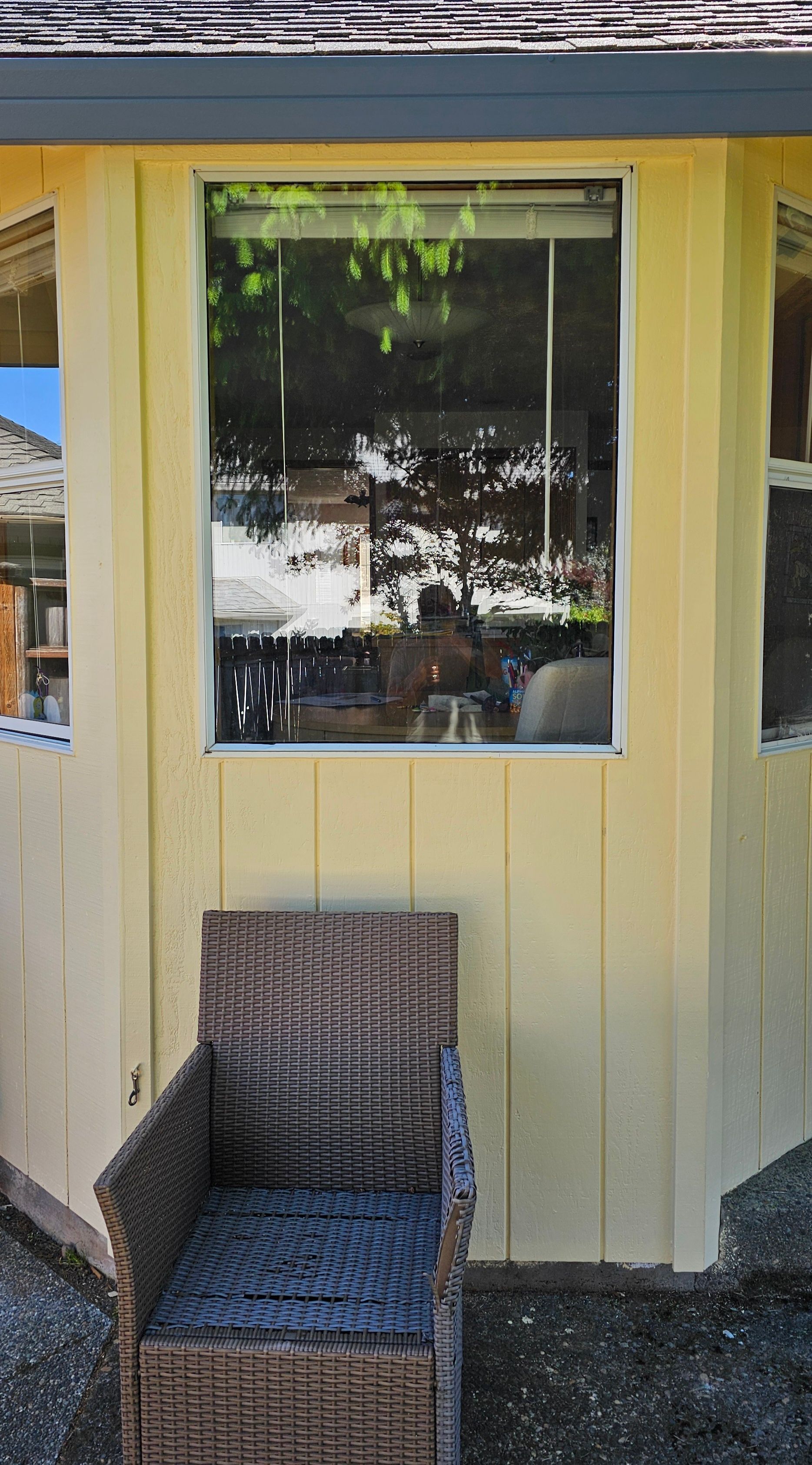 Wicker chair sits outside a yellow building with large window reflecting trees and interior.