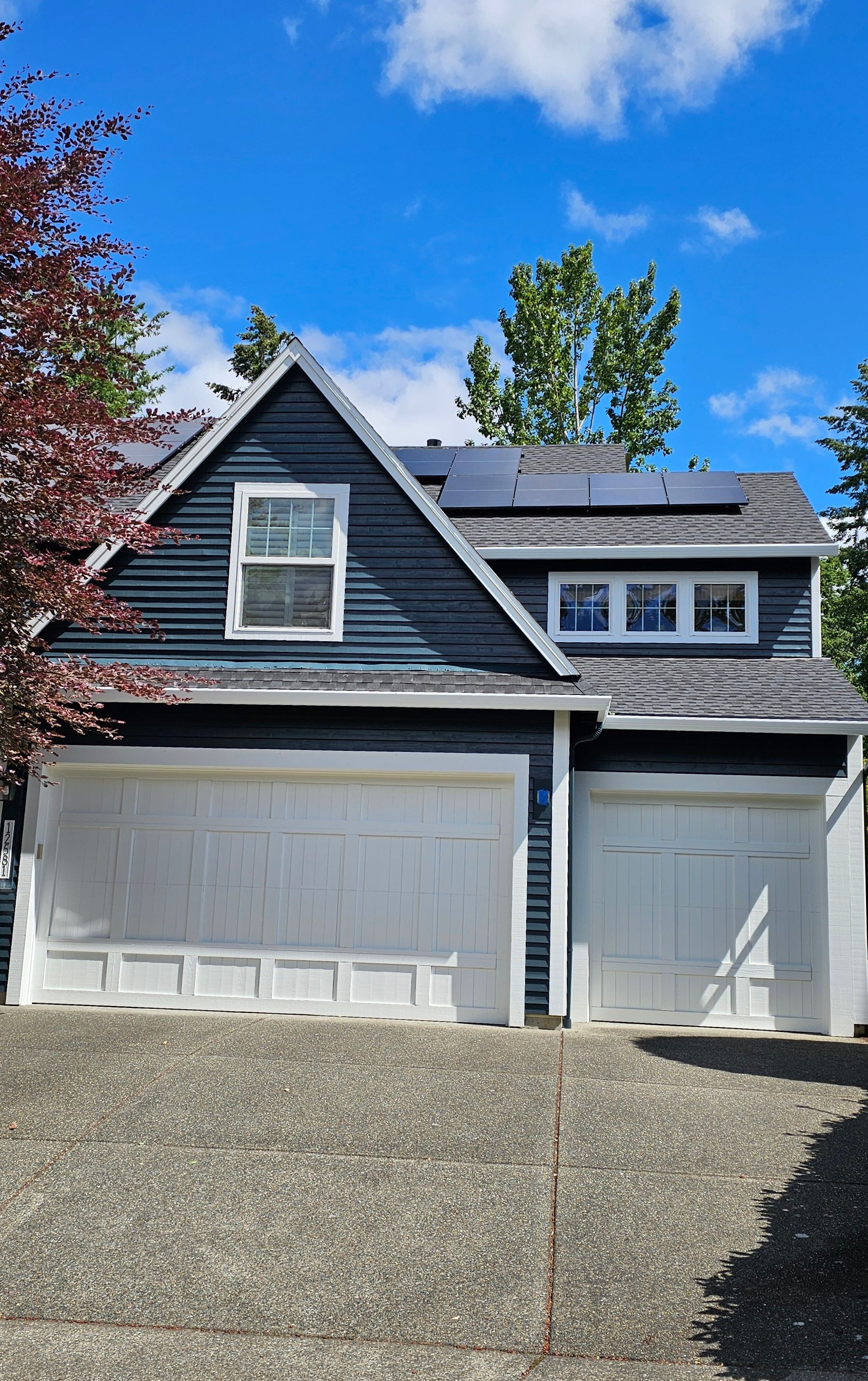 Two-story house with blue siding, white garage doors, and solar panels on roof against a blue sky.