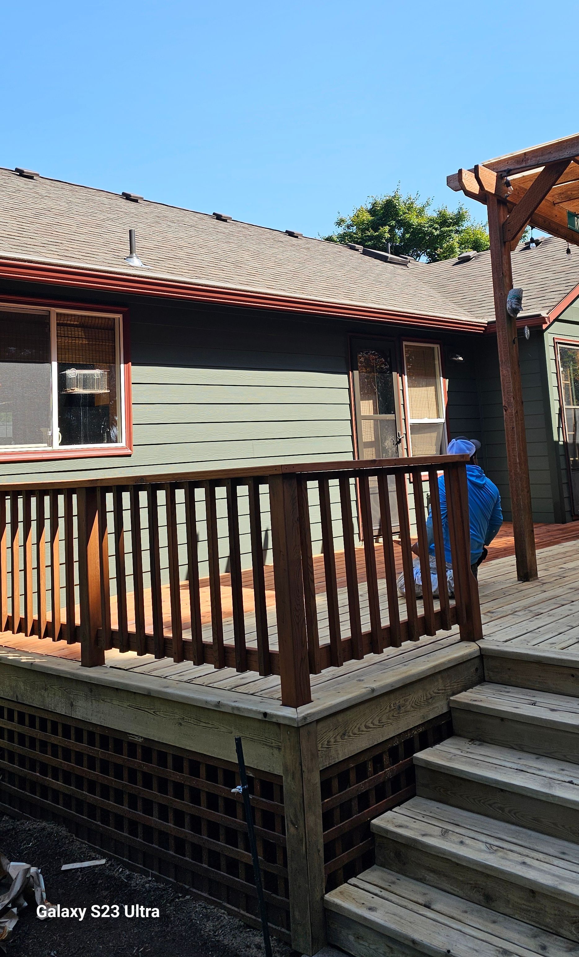 Deck with a brown railing, steps, and a pergola next to a green house with a red roof.