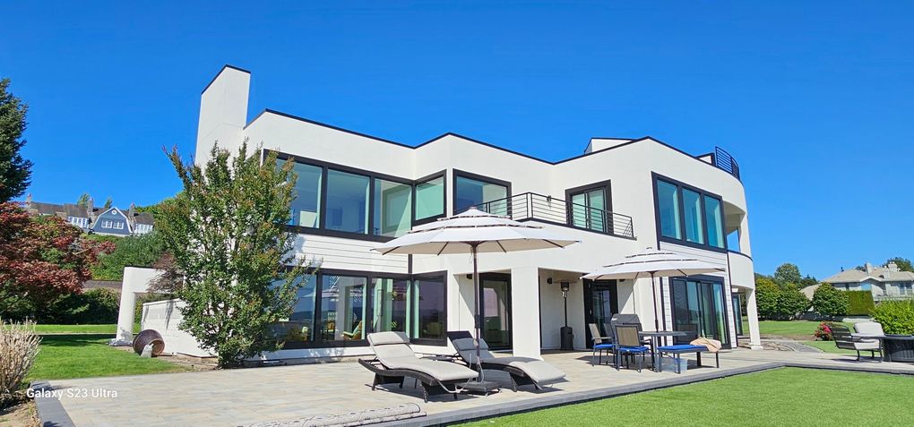 Modern white house with large windows, patio, lawn, and blue sky.