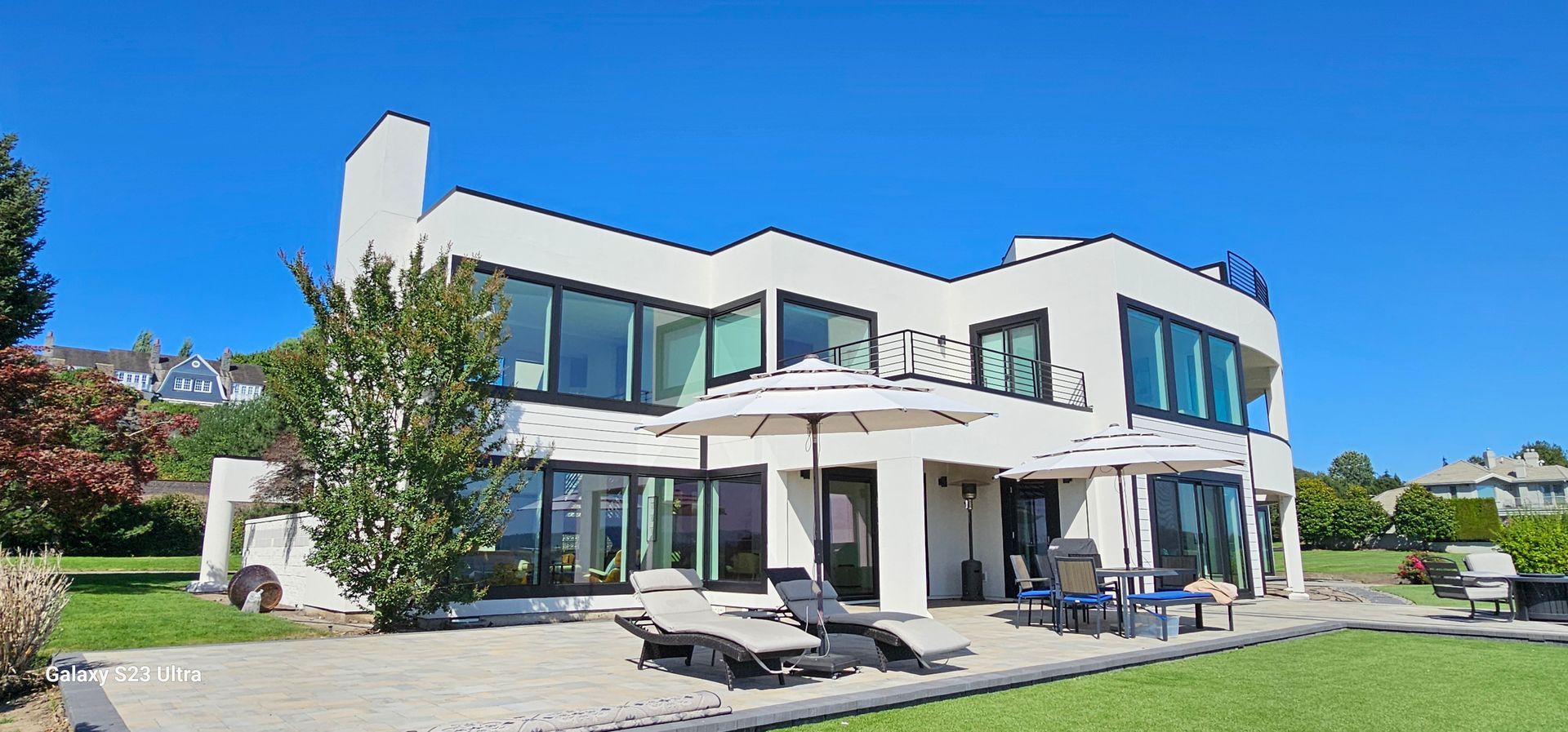Modern white house with large windows, patio, lawn, and blue sky.