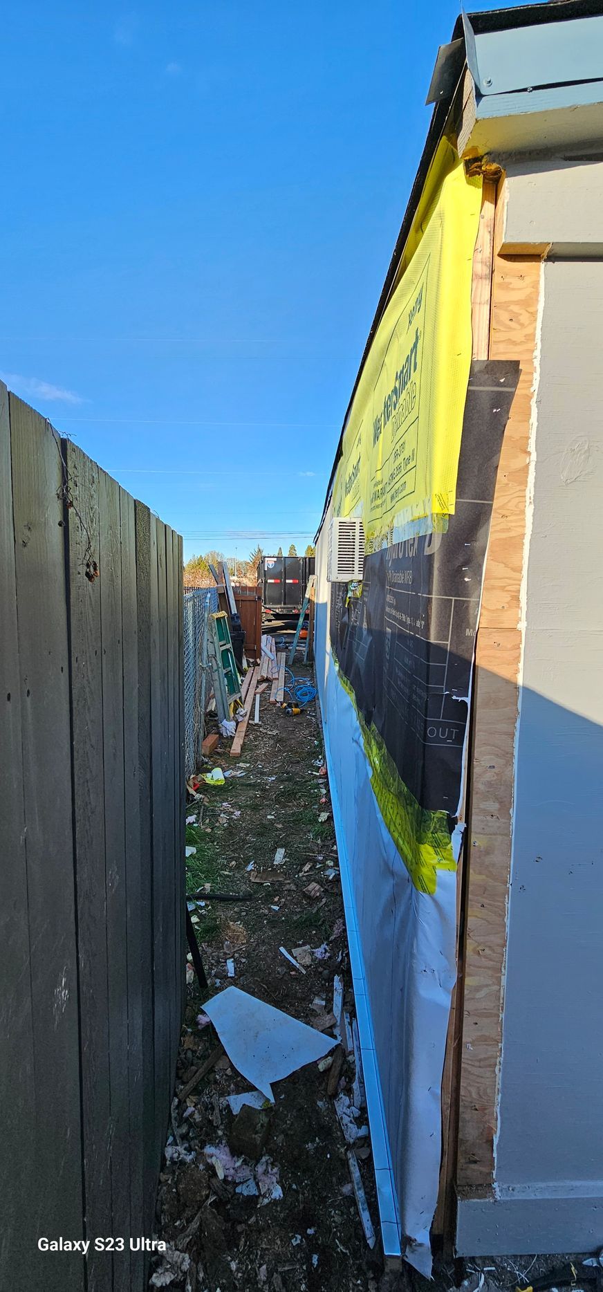 Narrow alley between a wooden fence and a building's exposed insulation and siding. Debris litters the ground.