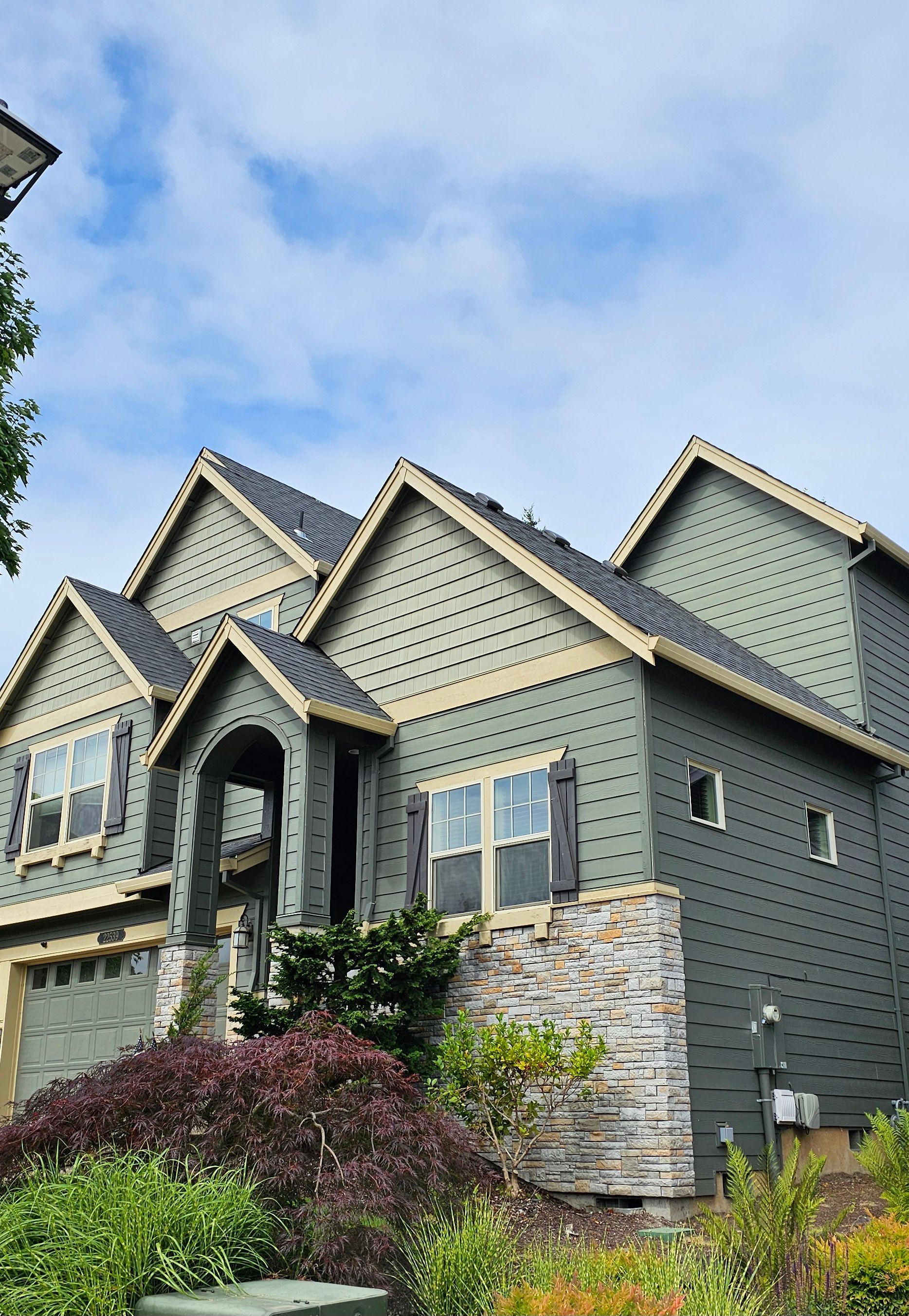 Green two-story house with stone accents, multiple gables, and landscaping under a cloudy sky.