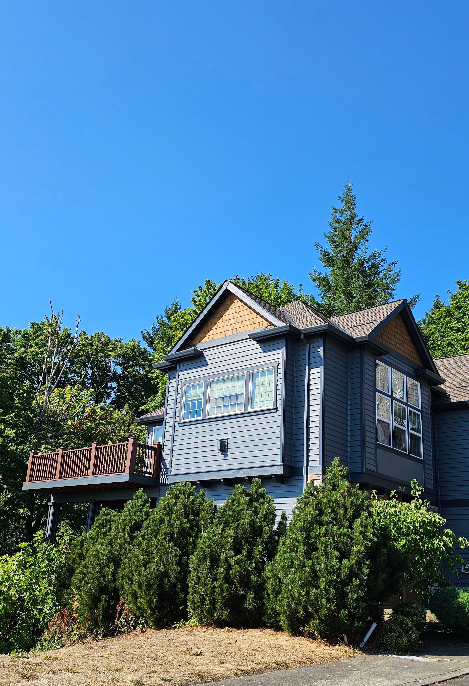 Blue-gray house with brown roof and trim, deck, and windows, surrounded by green trees under a bright blue sky.