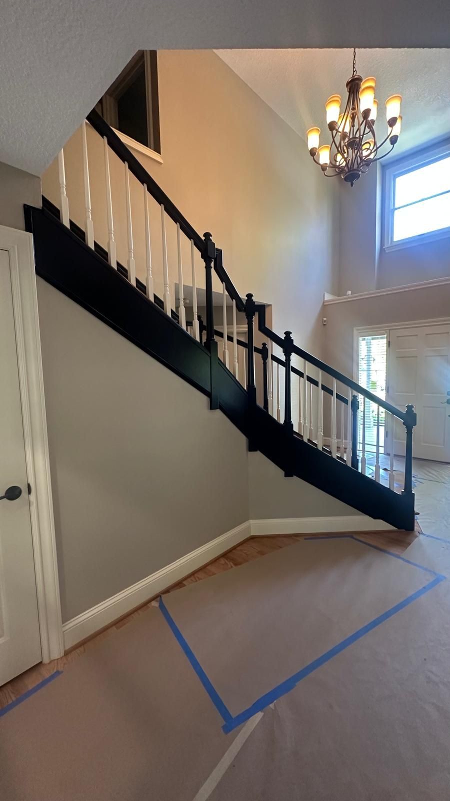 Staircase with black banister, white spindles, and tan walls. Hardwood floor covered with blue-taped paper.